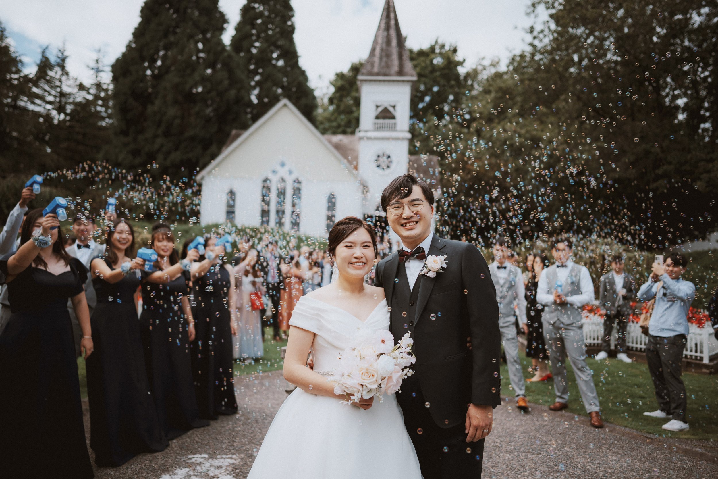Smiling bride and groom standing together with the chapel and guests behind them.