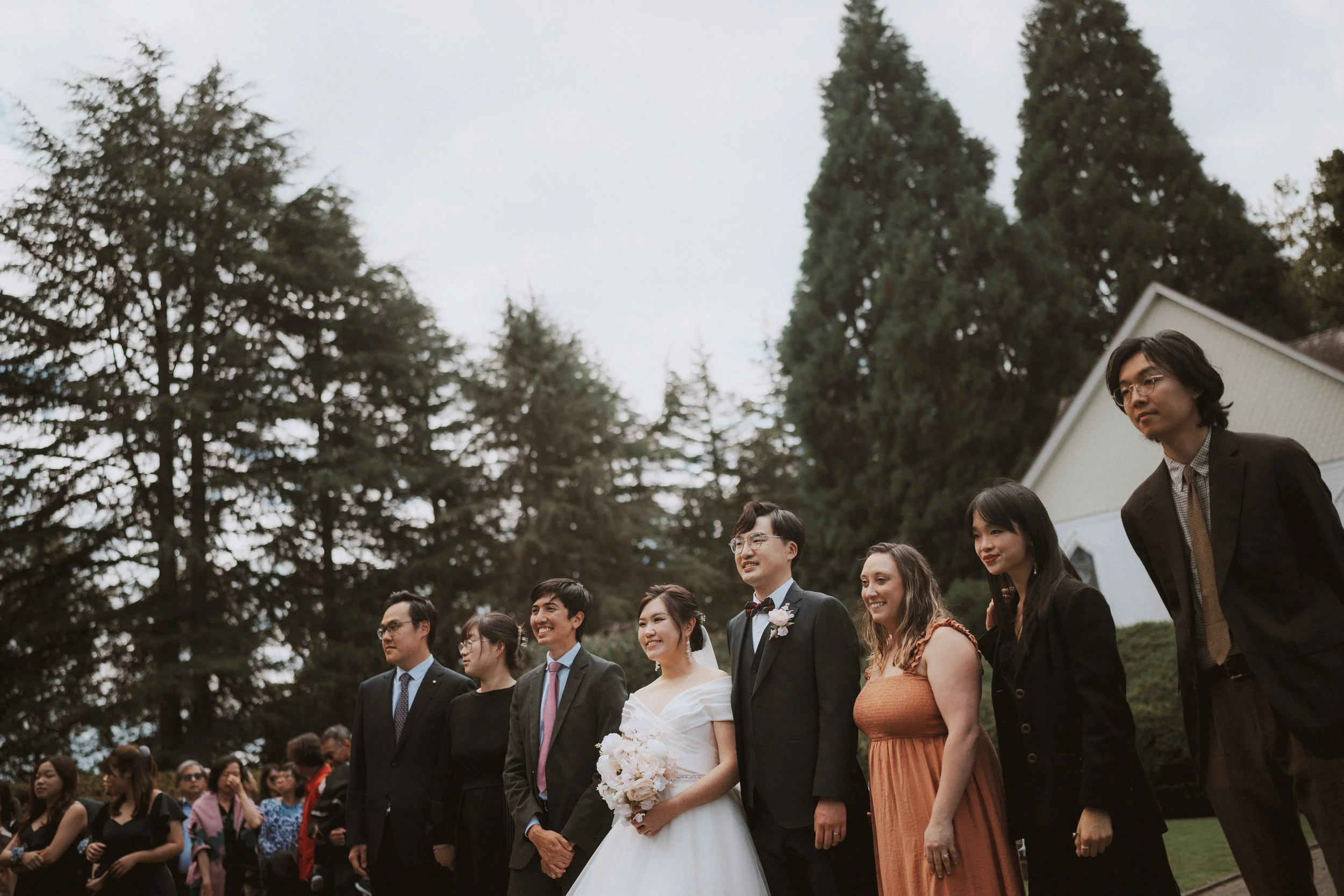 Couple posing with guests on the chapel lawn beneath tall trees.