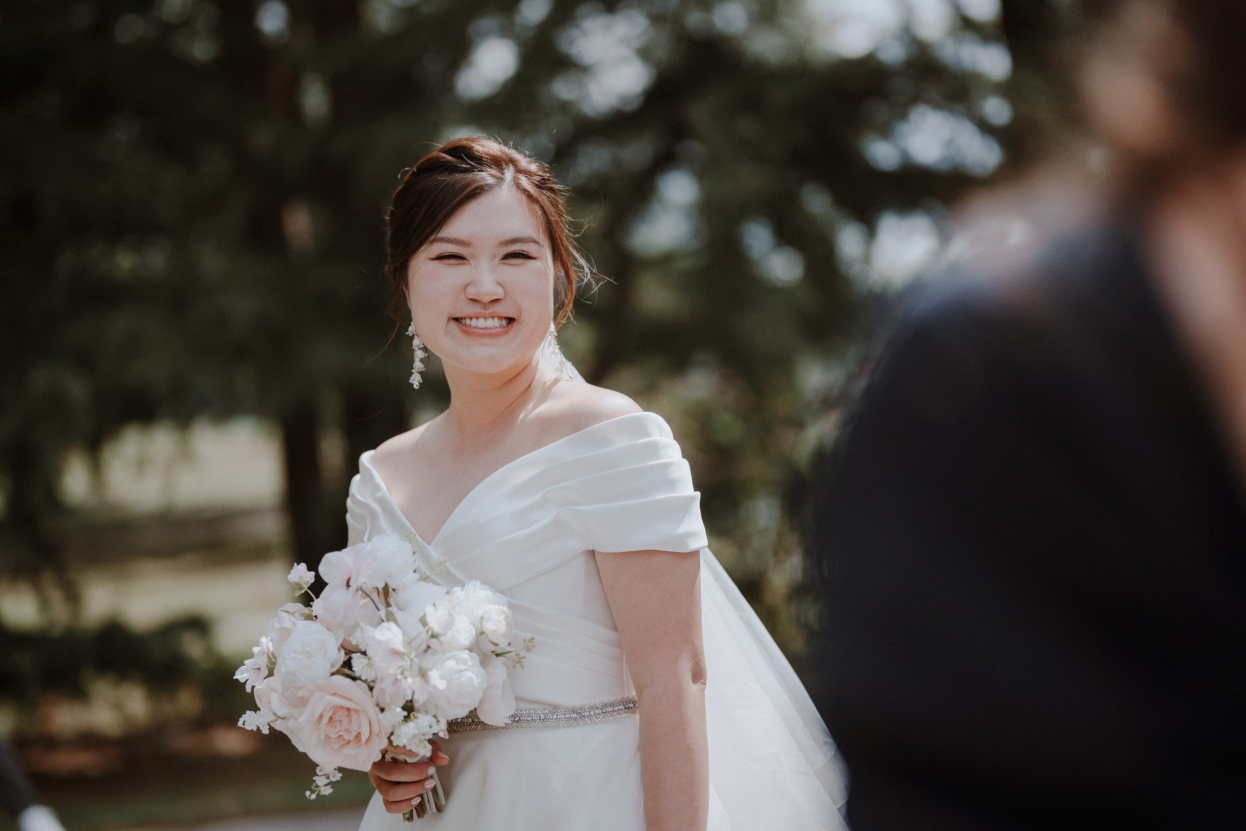 Bride smiling toward the groom while holding a pale wedding bouquet.