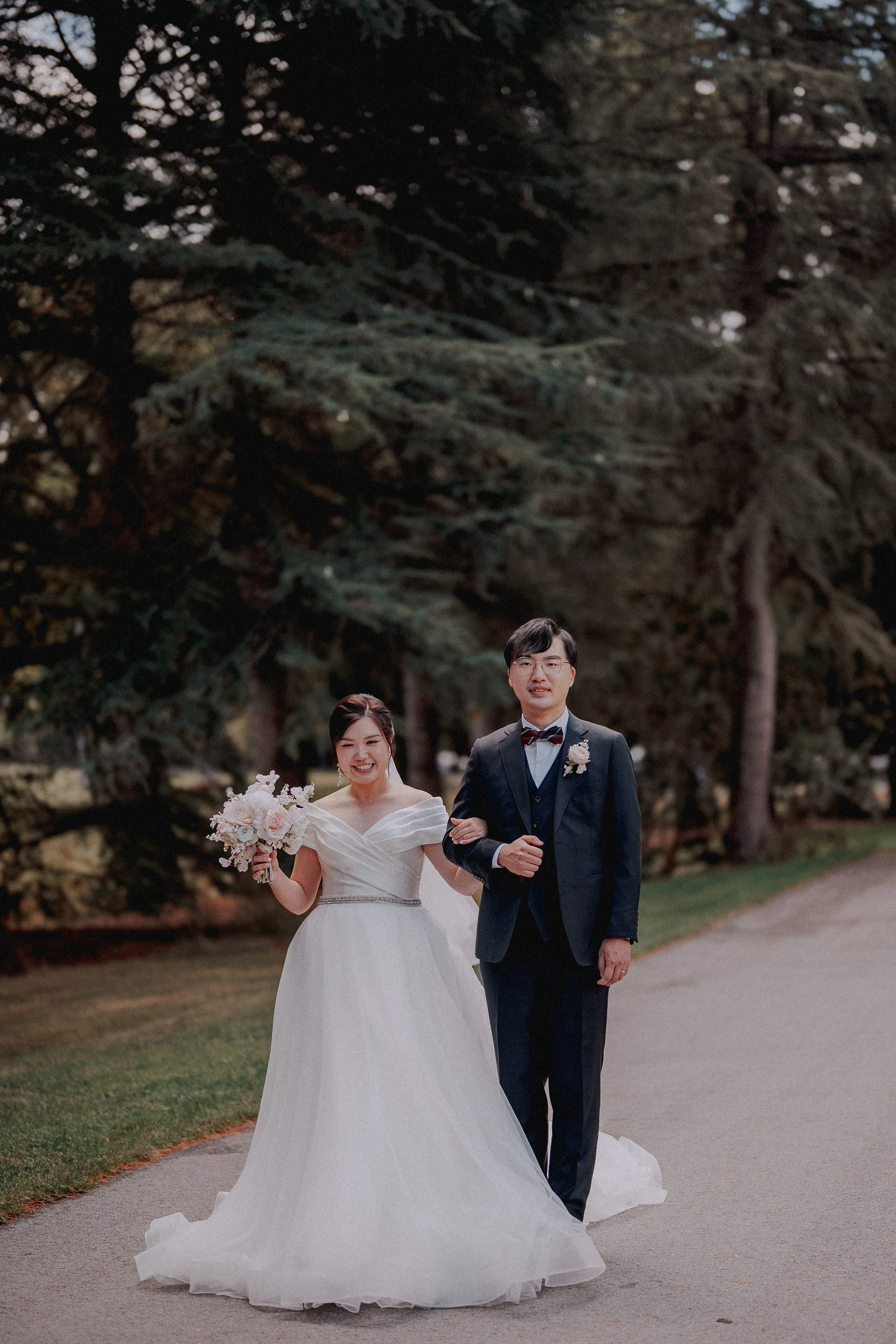 Bride and groom walking arm in arm along a tree-lined path.