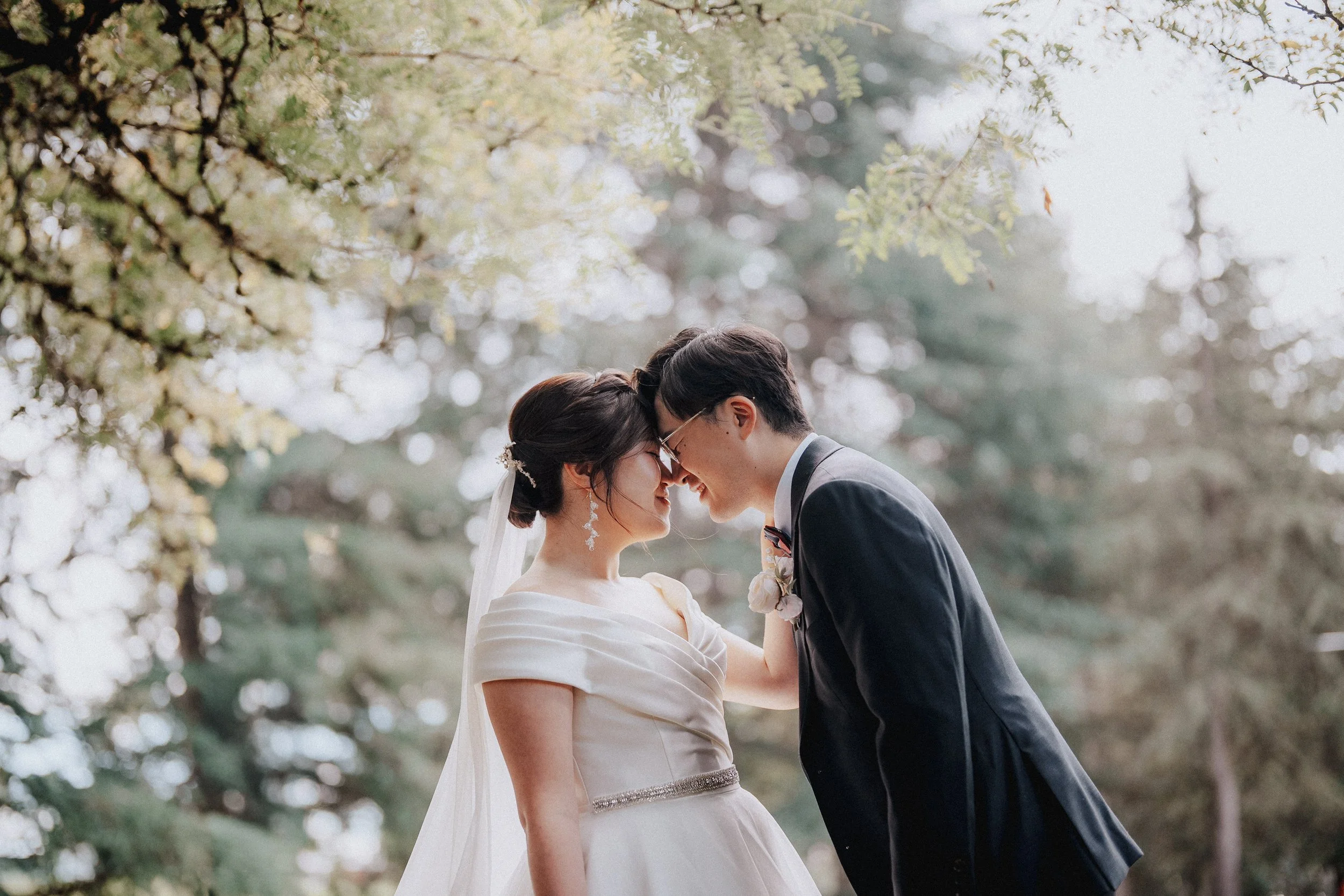 Bride and groom leaning together beneath sunlit branches.