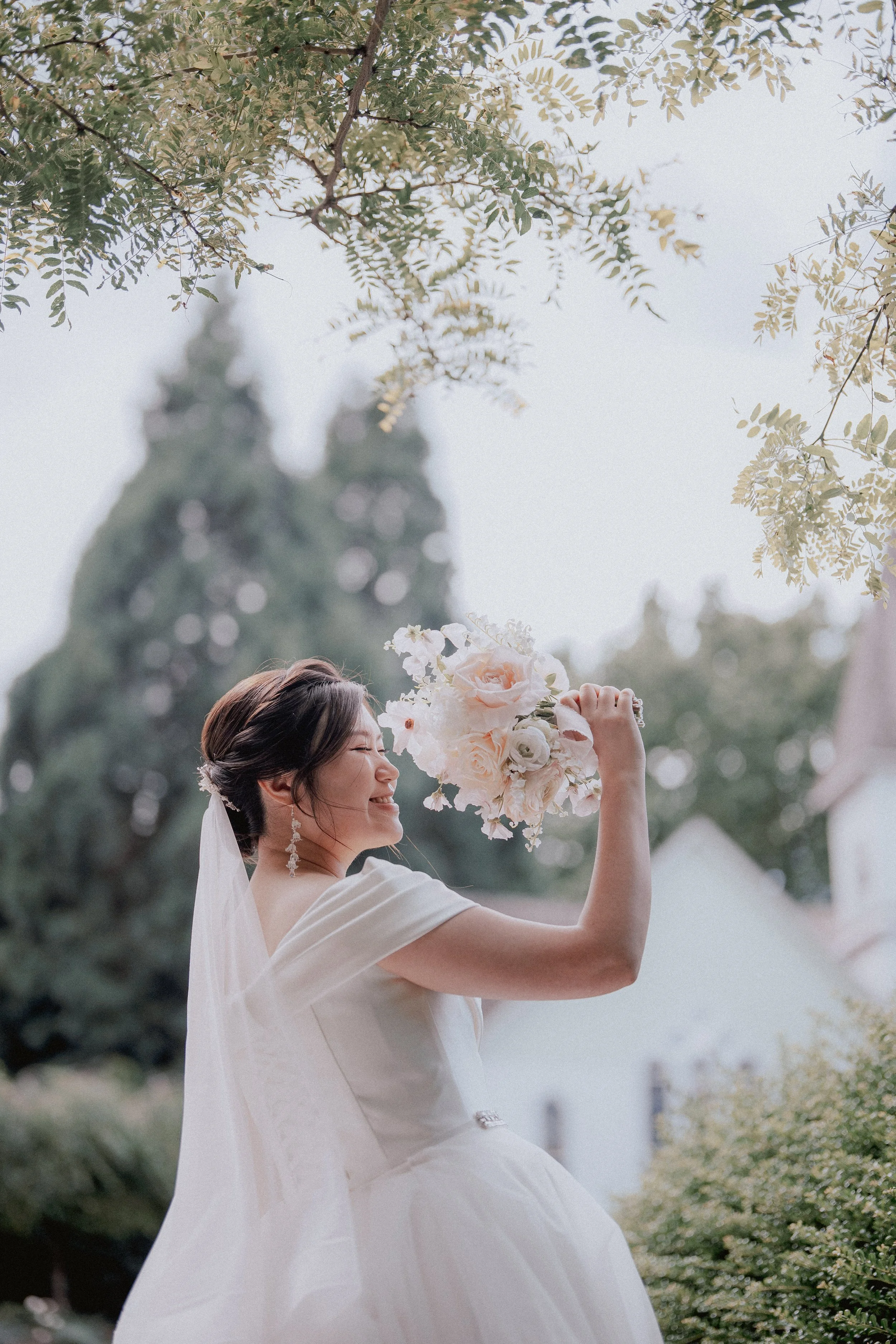 Bride raising her bouquet beneath the branches of a garden tree.