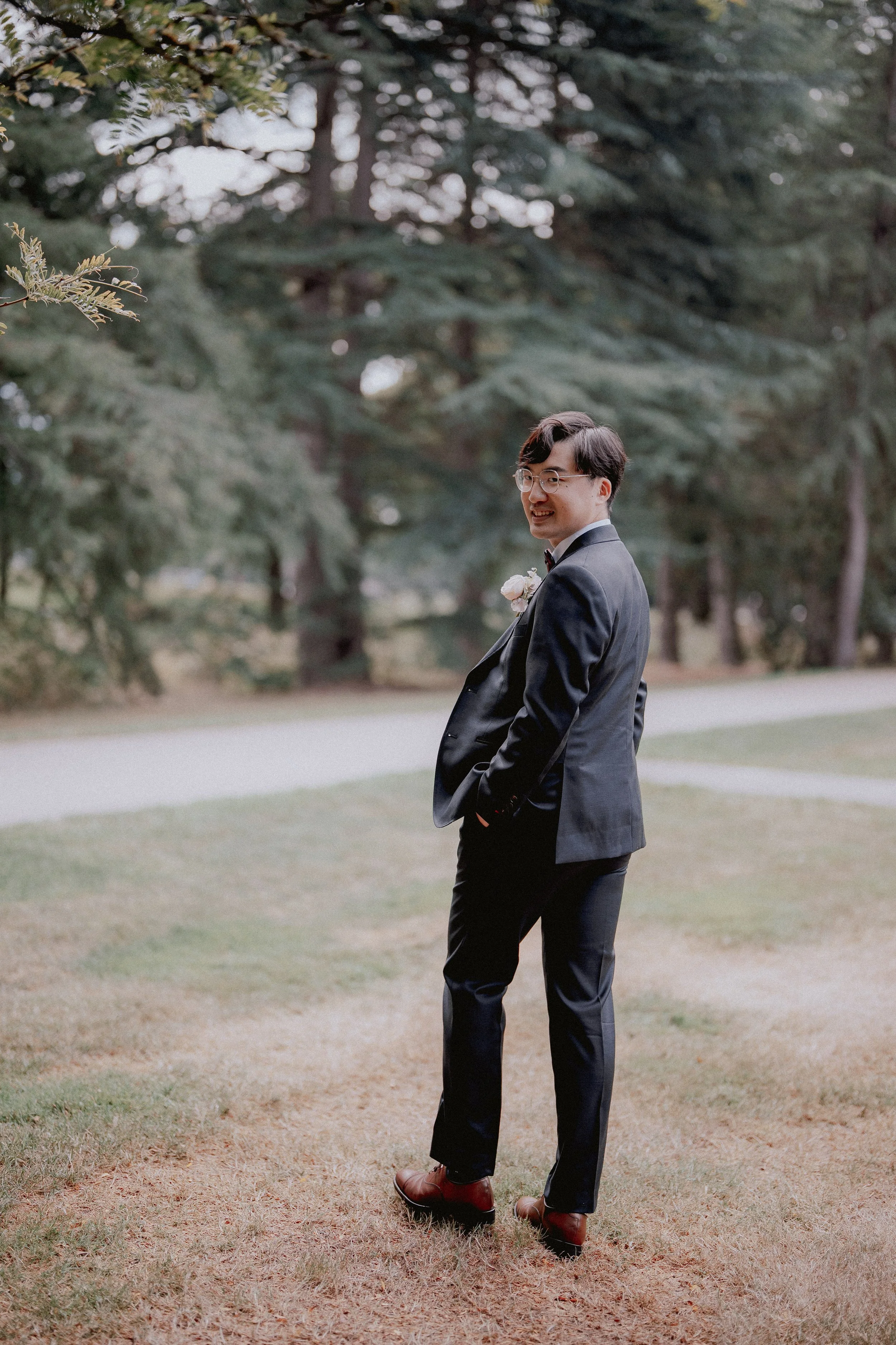 Outdoor portrait of the groom on the lawn beside tall trees.
