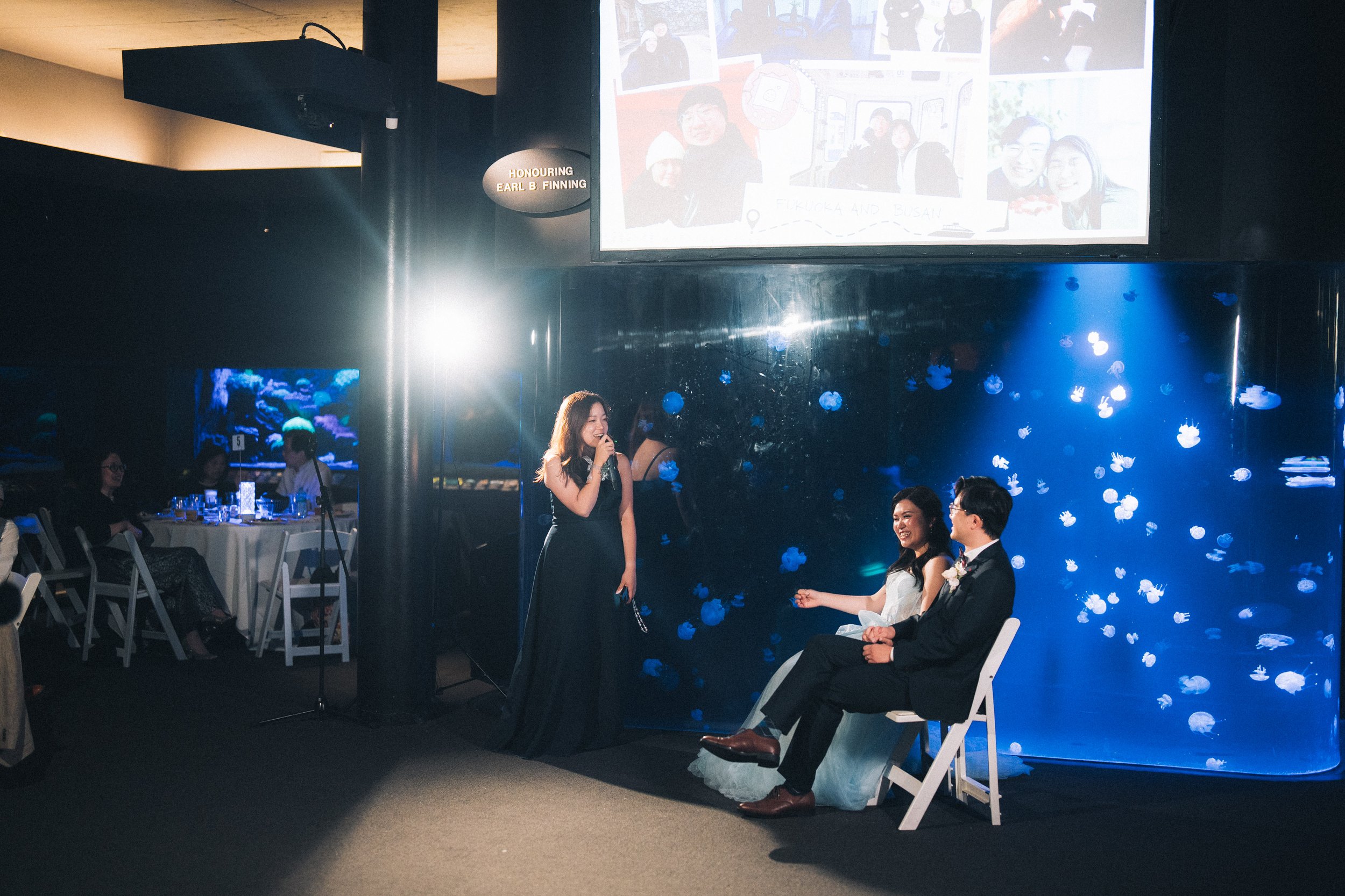 Guest speaking into a microphone beside the seated newlyweds.