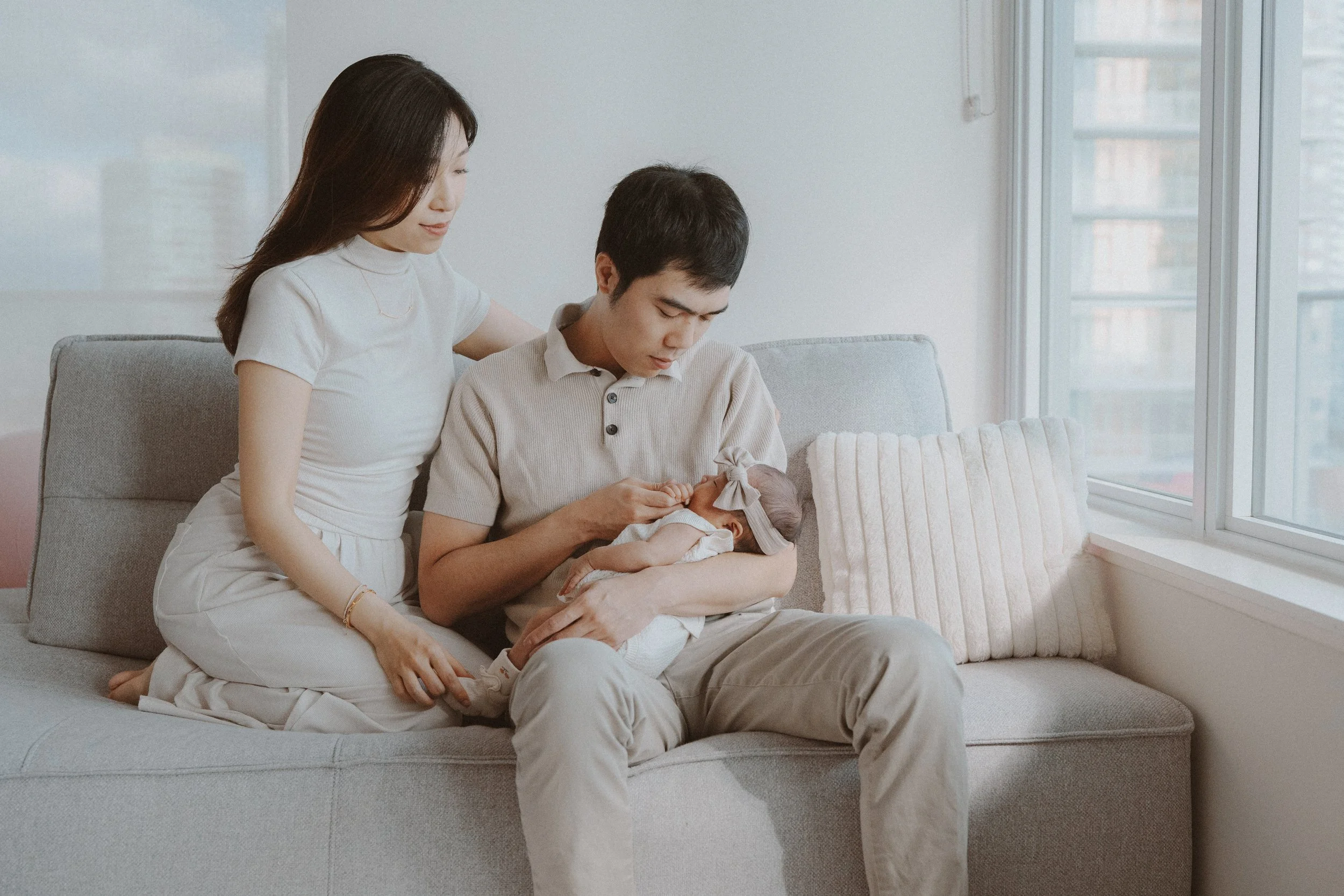 Wide family portrait of parents and newborn on a pale sofa beside a window.