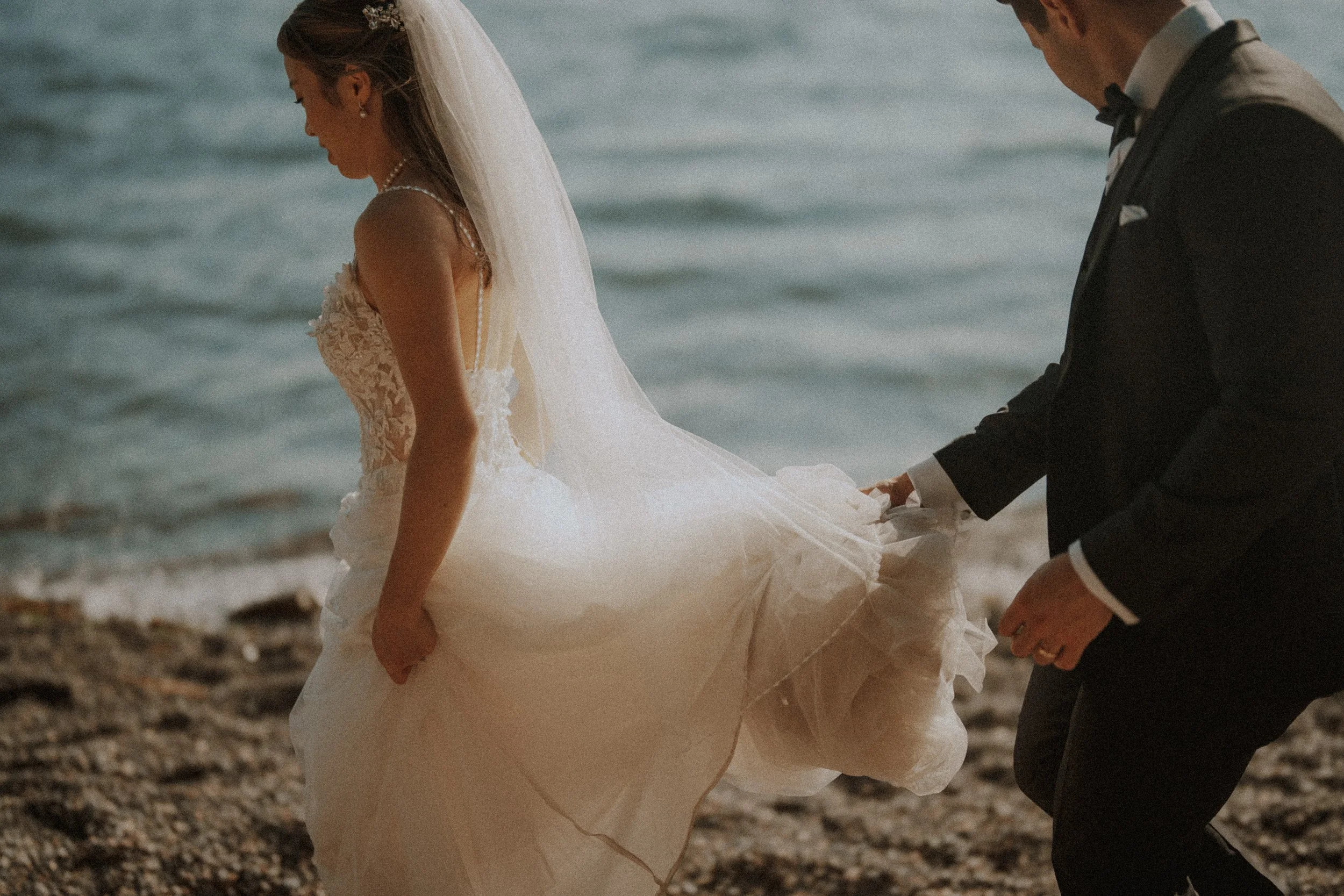  A tender detail shot of the groom gently lifting the bride's flowing wedding dress as they walk along the beach in  Whytecliff Park . 