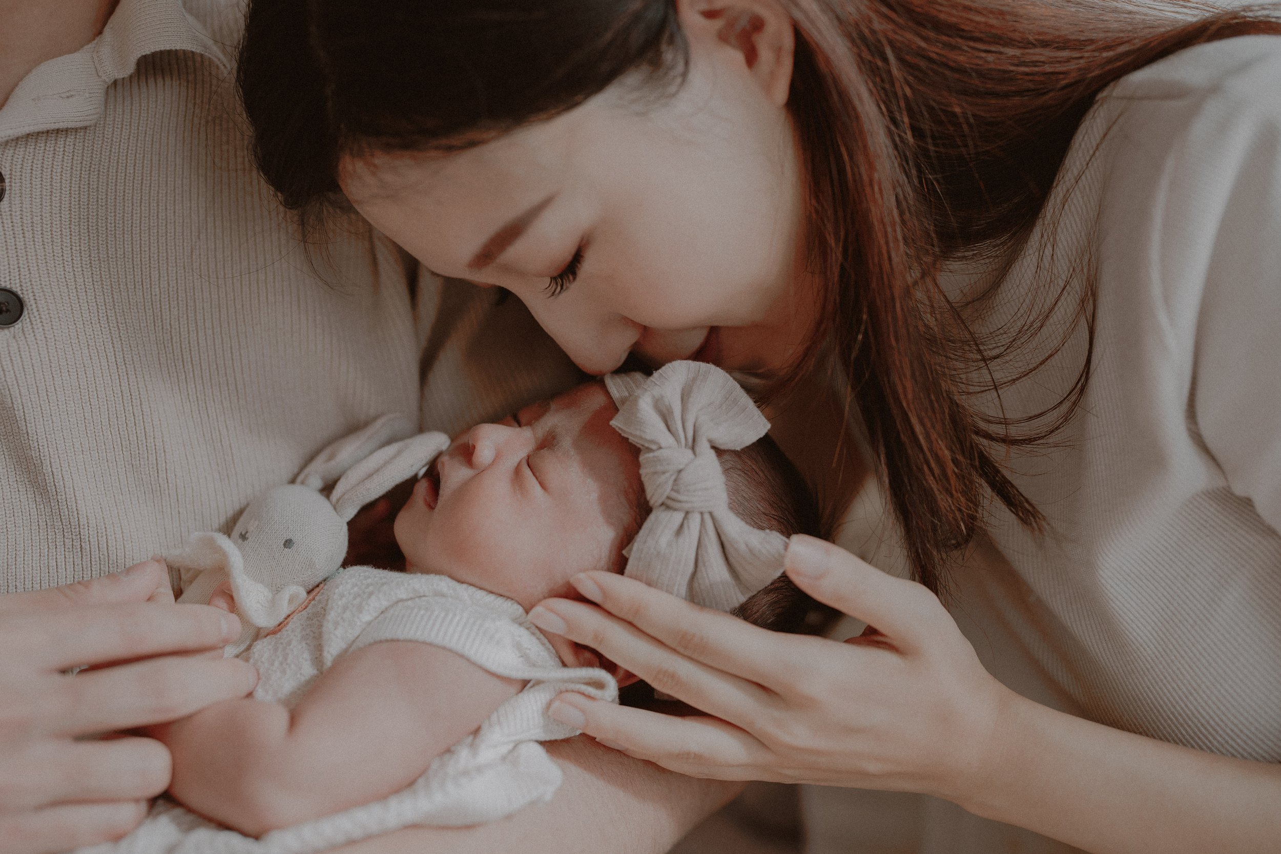  A heartwarming newborn photography session in Vancouver featuring Tata and Erik's baby girl. The infant is wearing a delicate bow headband and being cradled by her parents in a minimalist, earth-tone studio setting by 123 Cheese Production. 
