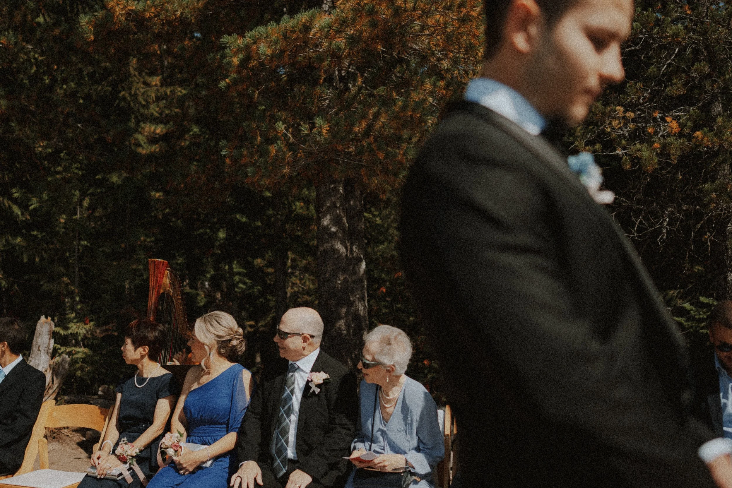  A powerful close-up of the groom's profile in the foreground, with guests attentively watching the outdoor wedding ceremony. Capturing the atmosphere and emotion at a  Sea to Sky mountain wedding  in BC. 