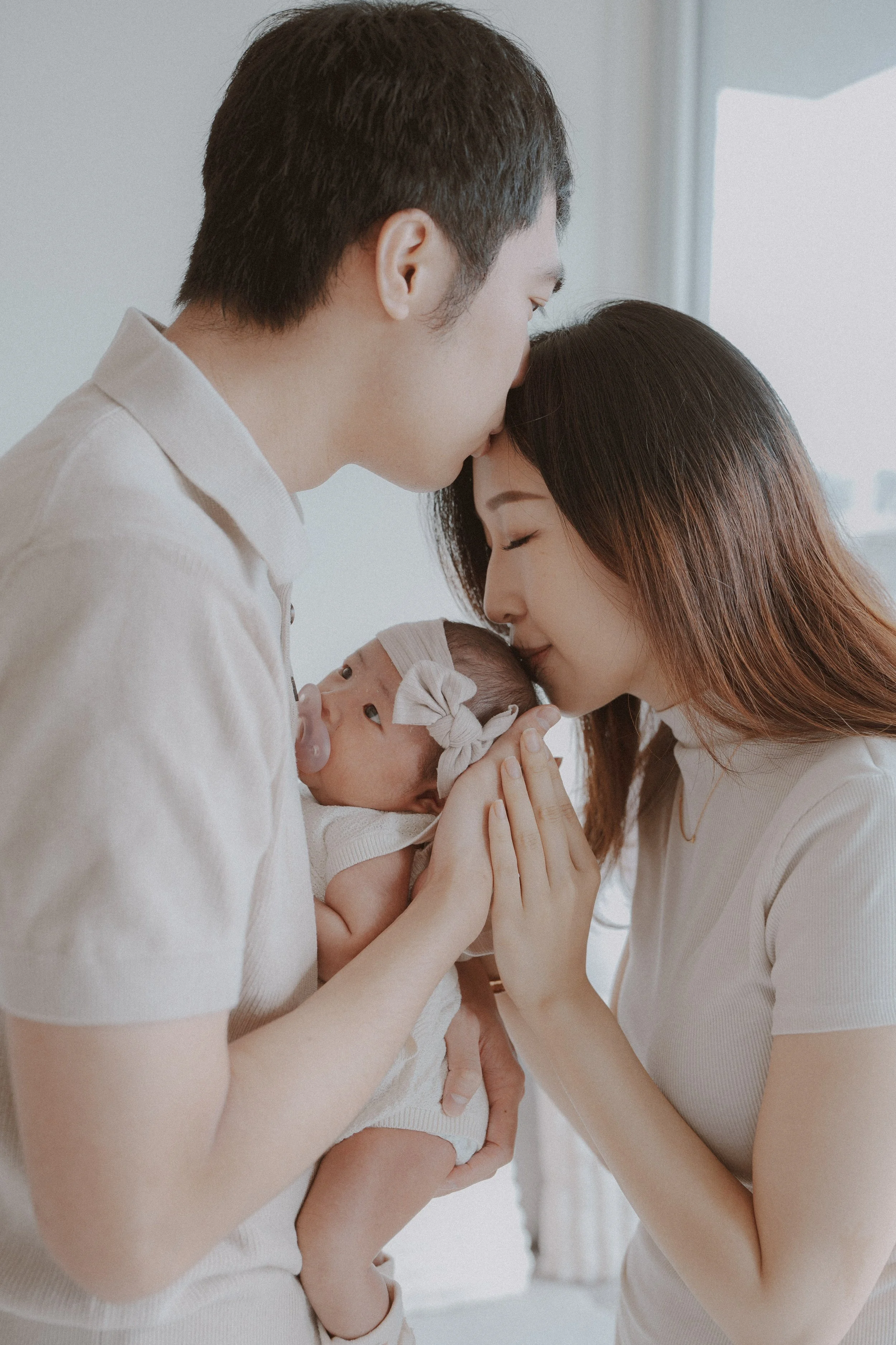 Father Lemon and mother Tata both kissing their newborn baby girls forehead while she opens her eyes featuring natural light with soft textures and warm hues in Vancouver.
