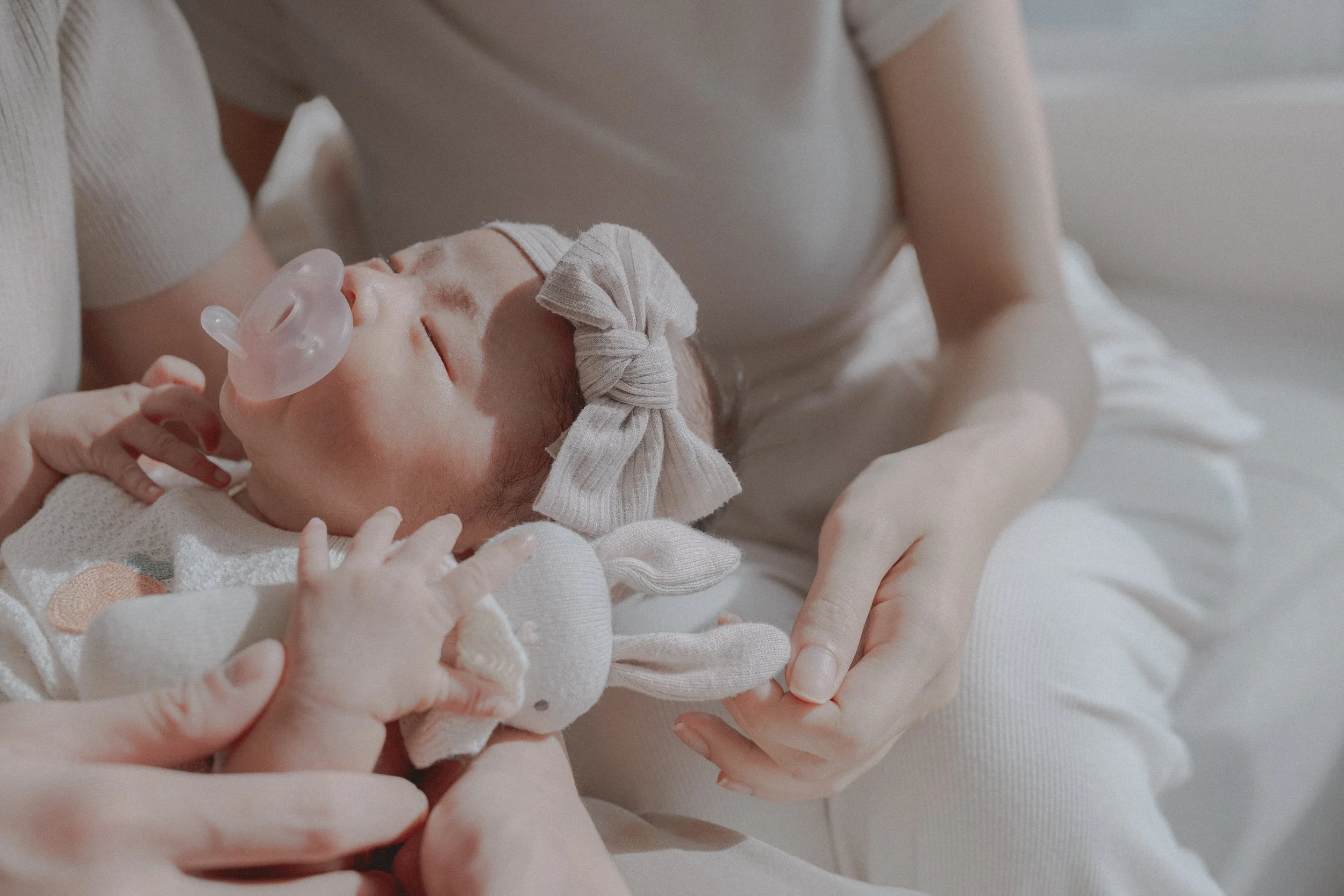 Newborn baby girl wearing a bow headband and holding a bunny toy while resting in mothers arms featuring natural light with soft textures and warm hues in Vancouver.