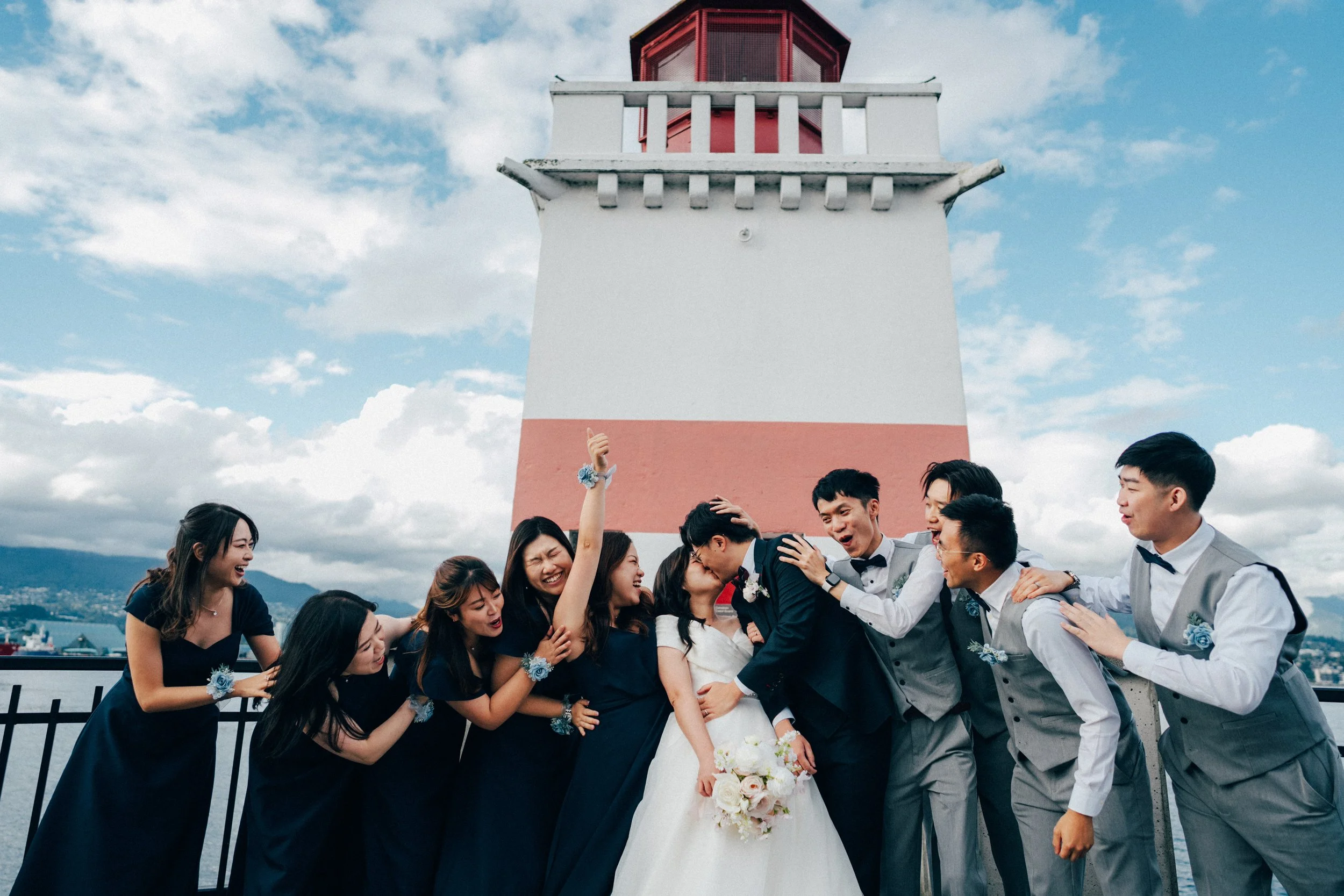  Pure fun and celebration! The bride and groom share a kiss, surrounded by their enthusiastic wedding party at the iconic  Stanley Park Lighthouse . 