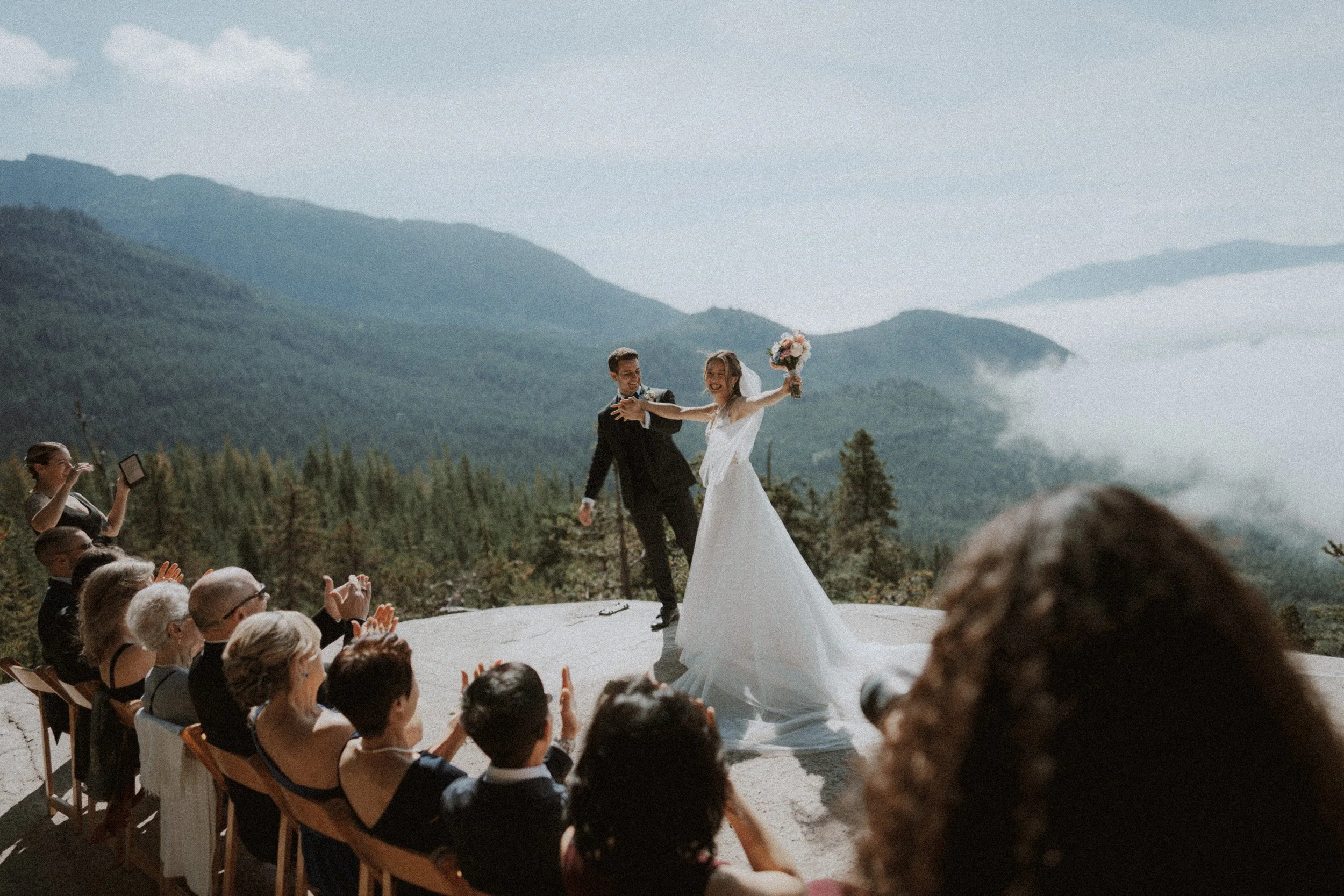  Pure celebration! The newlyweds cheer with their arms raised as guests applaud on the mountaintop. A joyful finish to their  BC mountain wedding . 