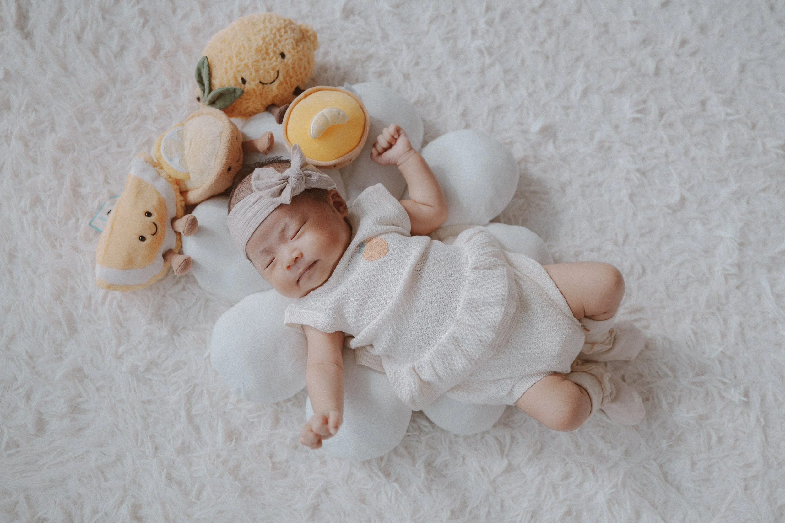 Newborn baby girl wearing a bow headband sleeping on a white cloud pillow surrounded by lemon plush toys using natural light to highlight soft textures and warm hues in Vancouver.