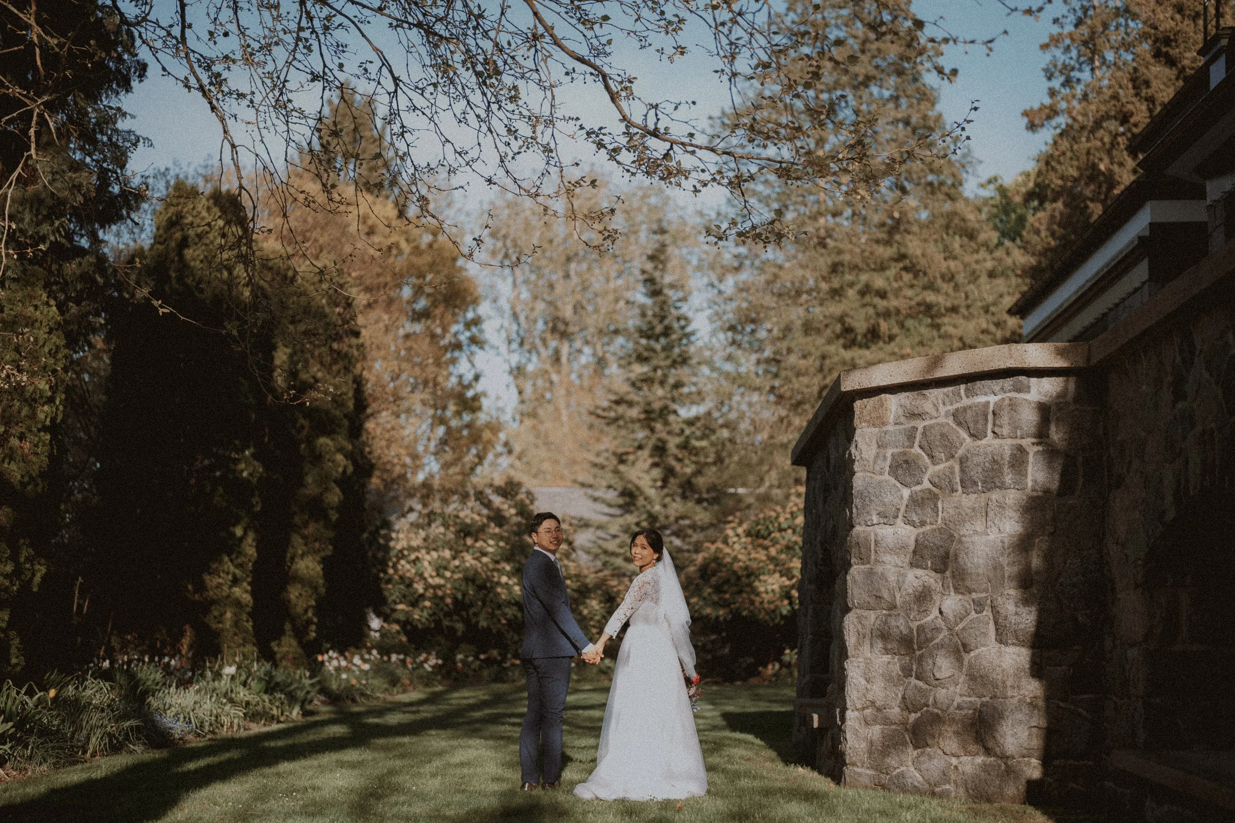  A romantic portrait of the couple holding hands, framed by the sun-drenched trees and the historic stone wall of the  Aberthau Mansion  grounds. 