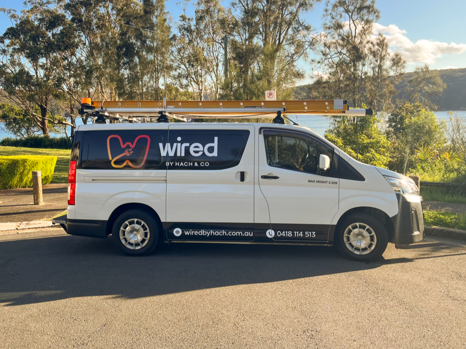 Company van for Wired by Hach & Co parked on a street with trees and water in the background, carrying a roof ladder.