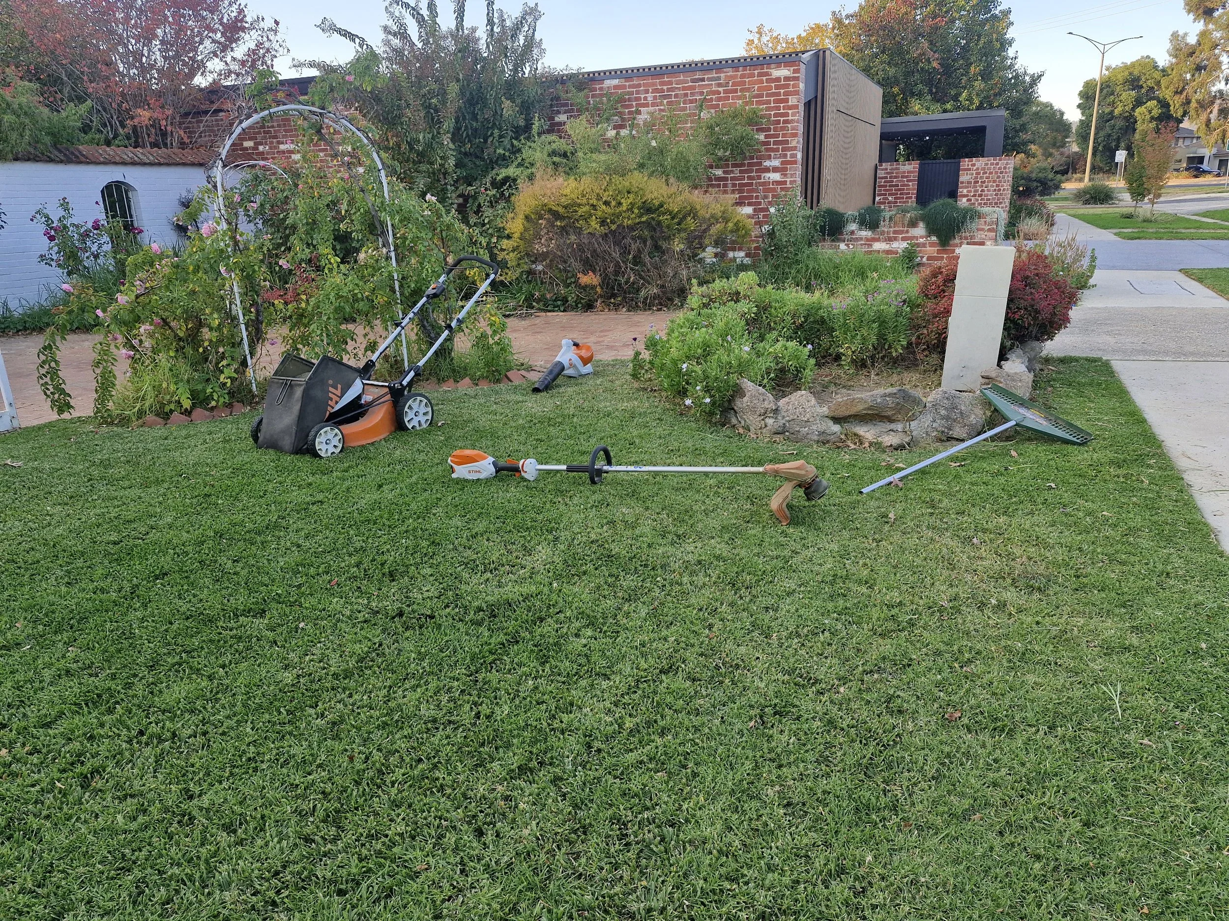 Lawn with gardening and yard work tools scattered, including a weed trimmer, rake, and leaf blower, near a flower bed with plants and rocks in front of a modern brick house.