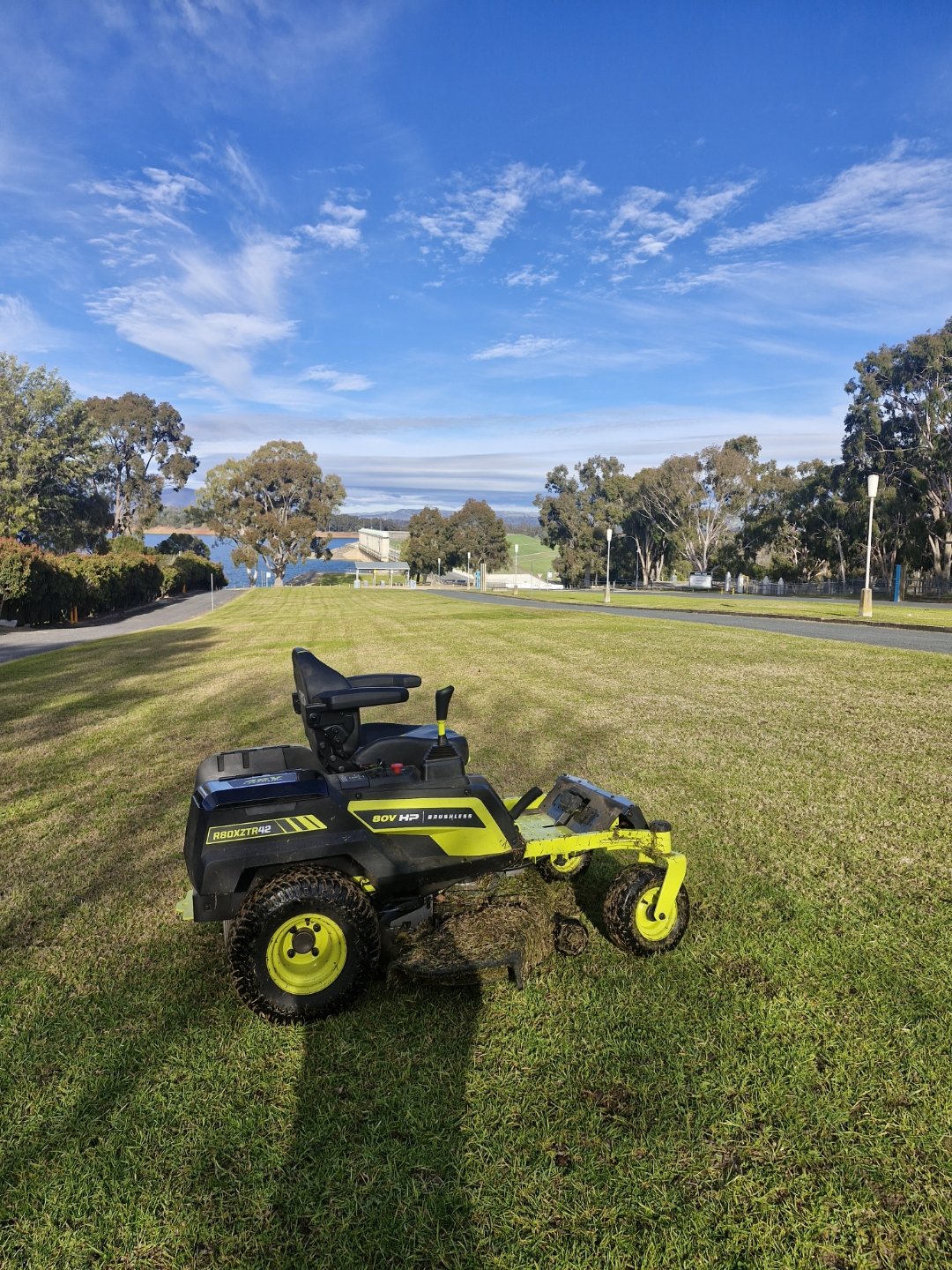A robotic lawn mower on a well-maintained grassy field under a blue sky with scattered clouds, surrounded by trees and a park area.