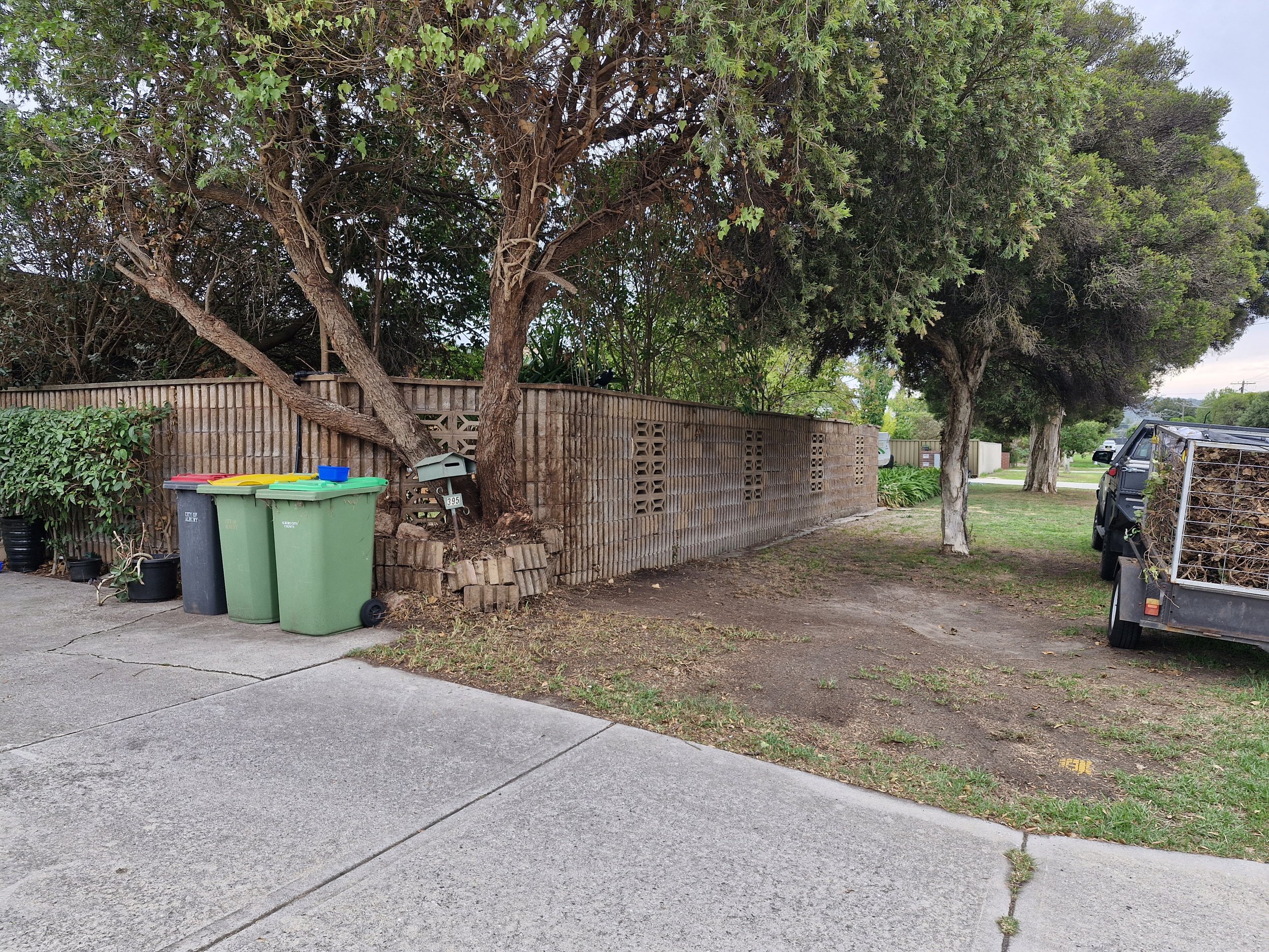 Residential backyard with trees, a gravel pathway, a wooden fence, trash and recycling bins, a black trash can, a small trailer filled with yard debris, and a mailbox.