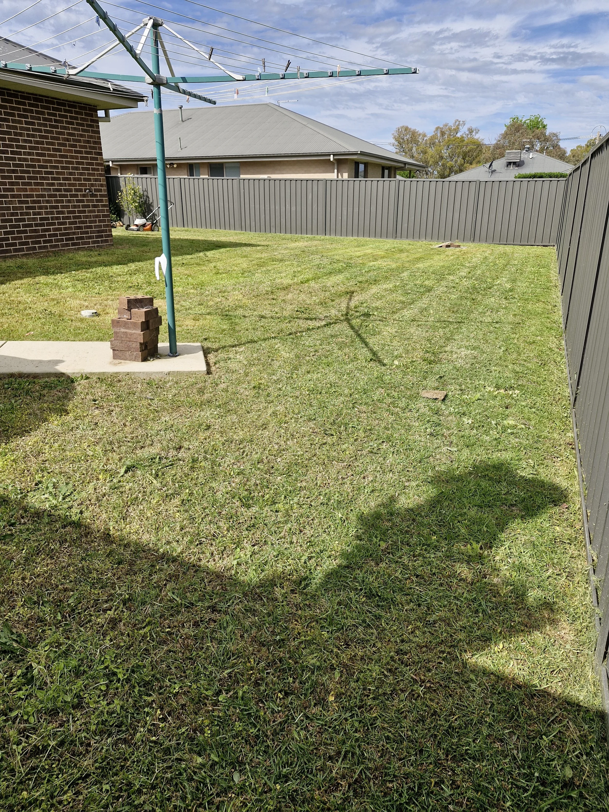A backyard with a rotary clothesline, a brick and concrete patio, a gray wood fence, and neighboring houses. The sky is partly cloudy.