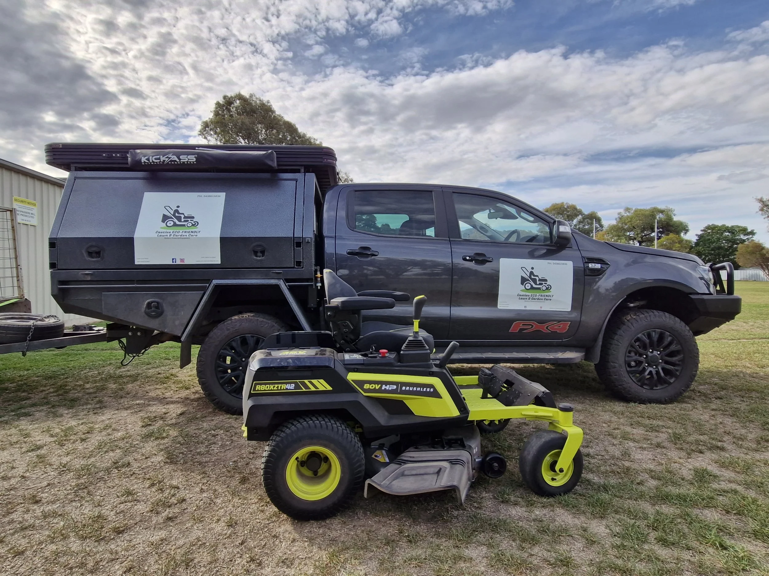 Black pickup truck with a lawn care logo and signage, parked on grass, with a yellow and black zero-turn lawn mower in front of it, under a partly cloudy sky.