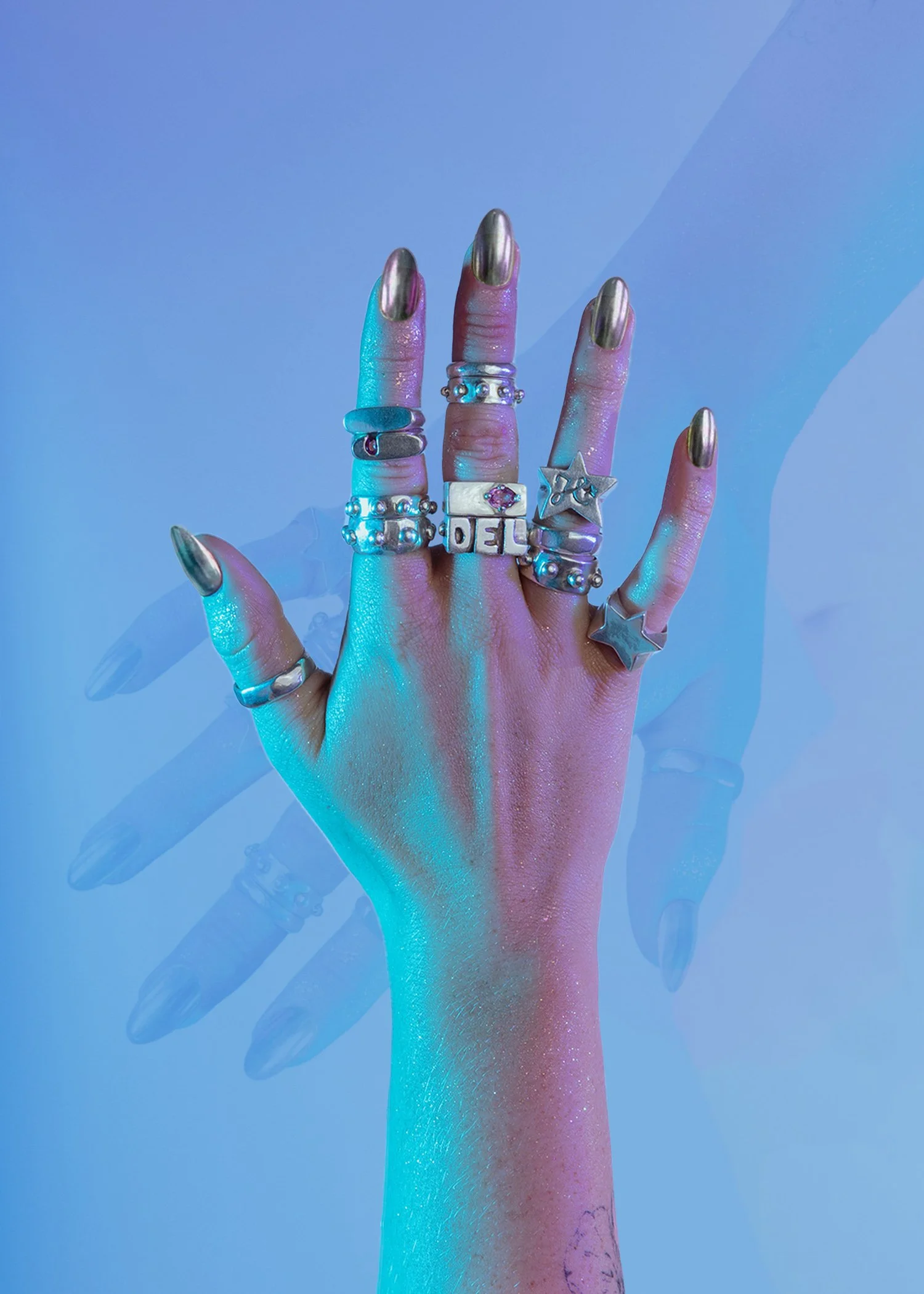 A hand with nails painted metallic gold, wearing multiple chunky 925 silver rings, is raised against a blue background with colored lighting effects.