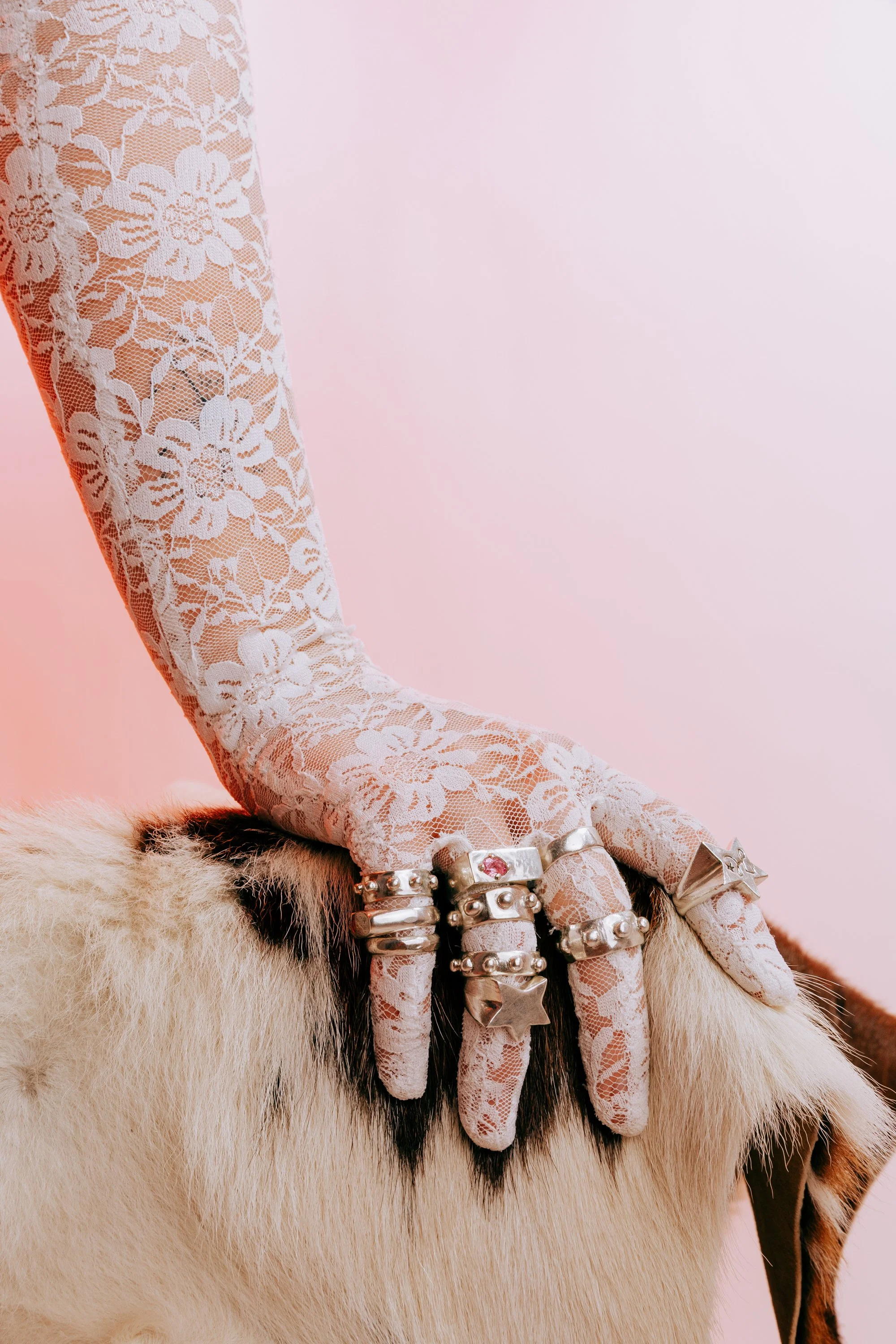 Close-up of a hand wearing white lace gloves and chunky, handmade 925 silver rings, resting on cow-hide with a pink background.