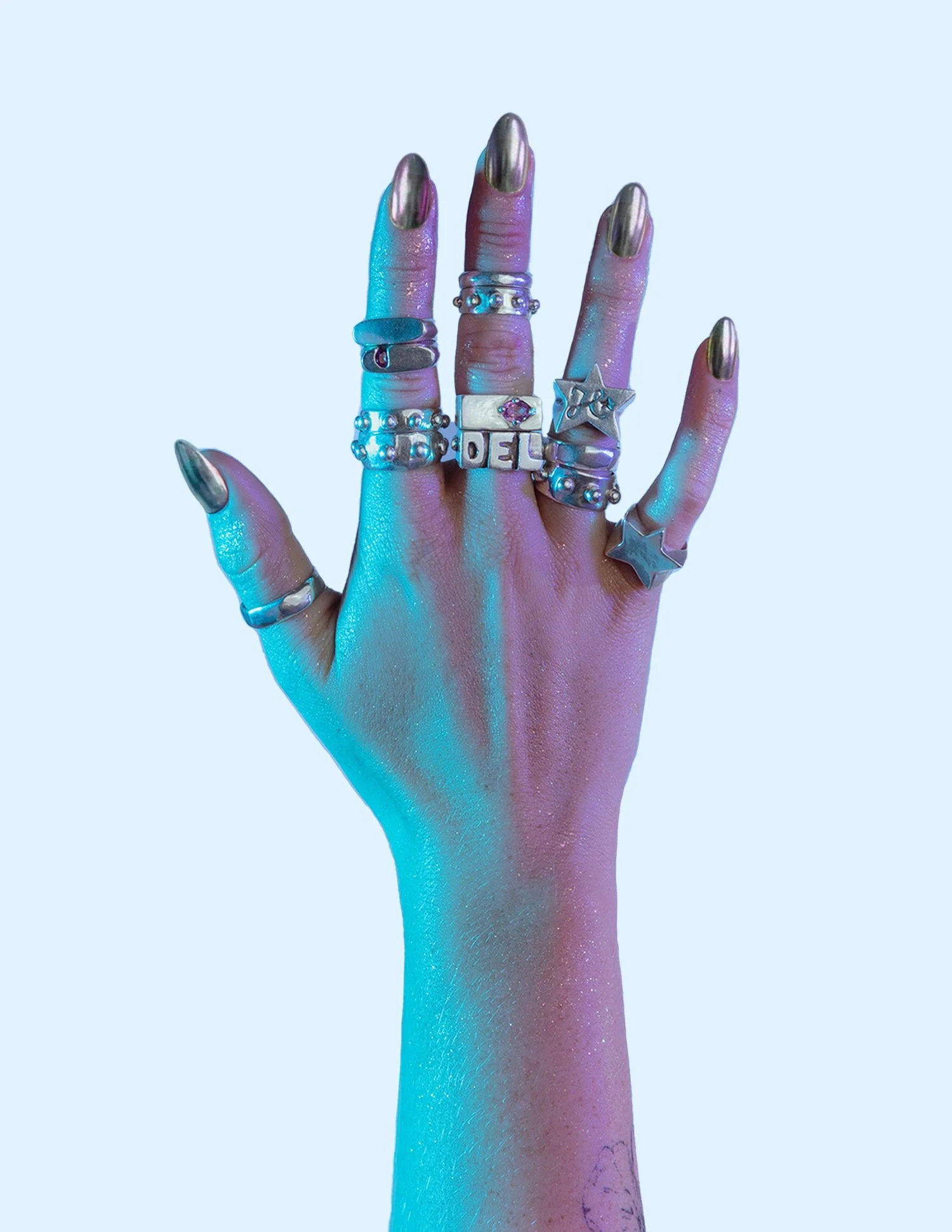 A human hand with metallic gold nail polish, wearing multiple rings on each finger, set against a light blue background with colorful lighting reflections.