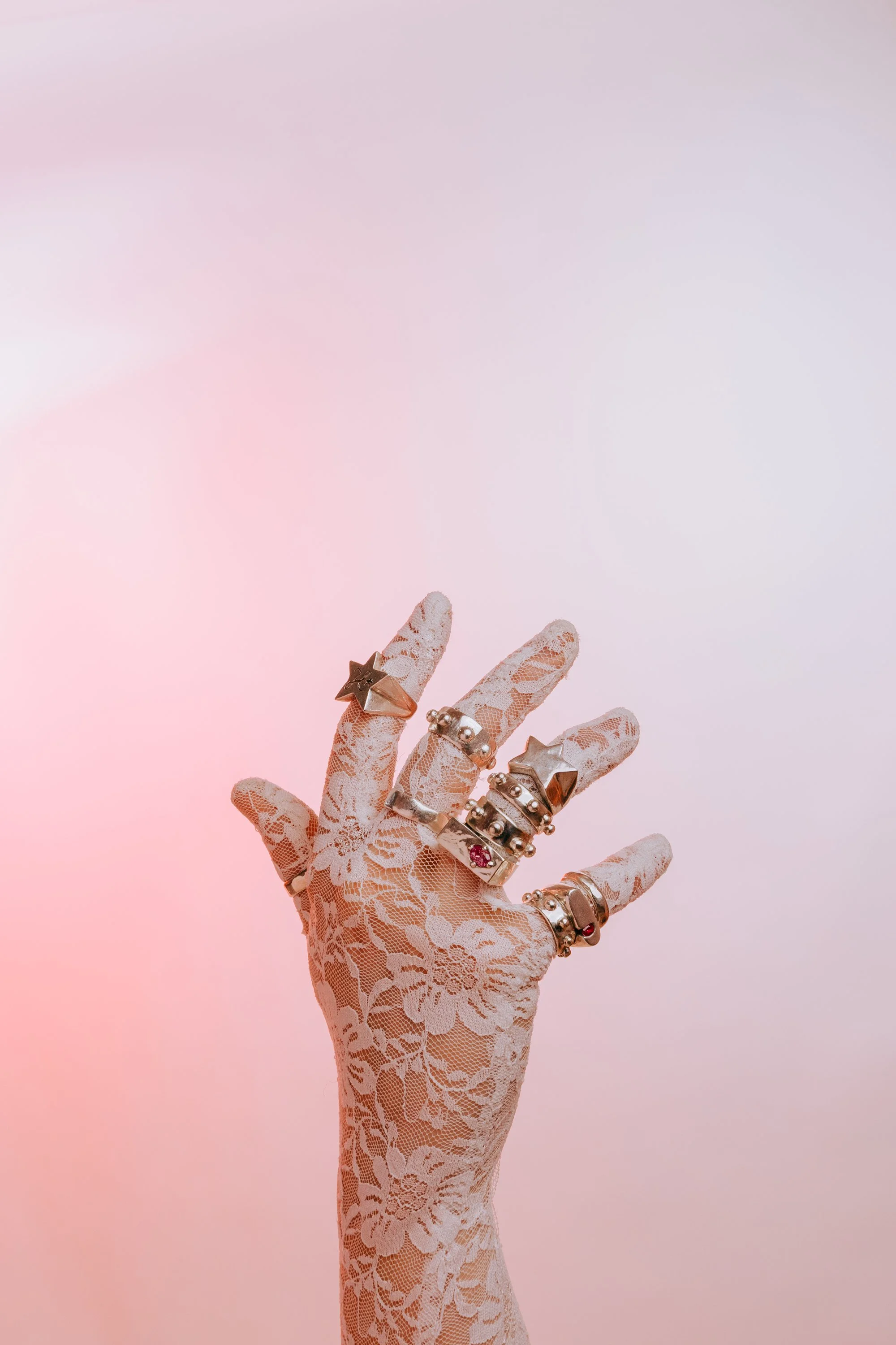 A hand wearing a lace glove with multiple rings, including the star ring, the bobble ring and pink gem 925 silver rings, against a pink and purple gradient background.