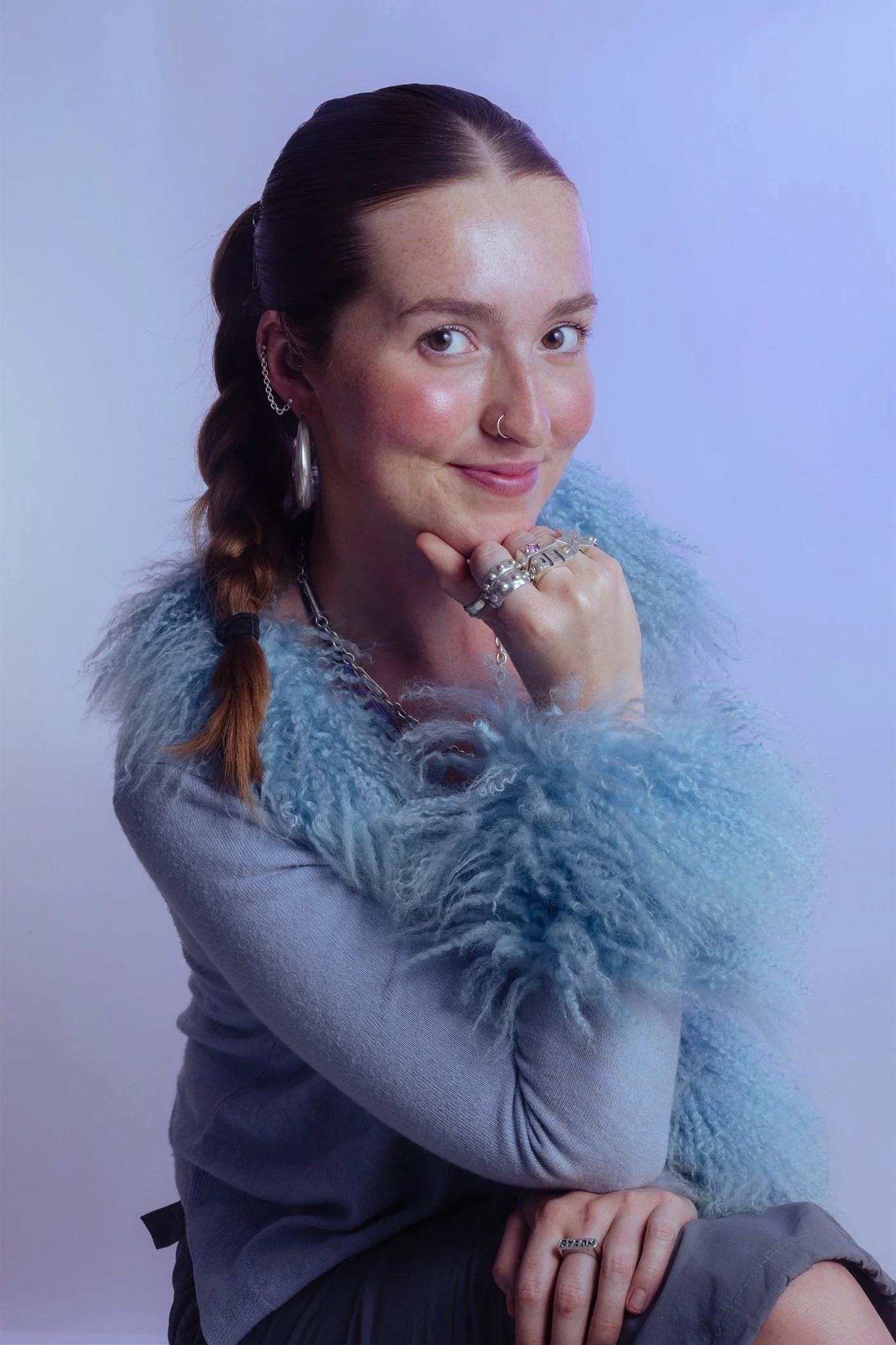 A woman with long brown hair styled in a braid, wearing silver jewelry including rings, earrings, and a nose ring, smiling with her chin resting on her hand. She is dressed in a light blue fluffy sweater and gray pants, set against a soft, gradient background.