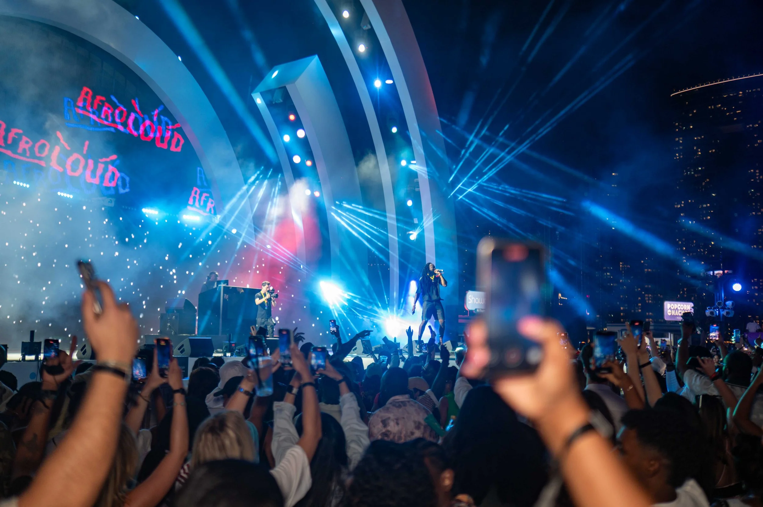 A concert stage with blue and white lights, a performer standing with a microphone, and a crowd of people holding up phones to record the event, set in an outdoor urban area at night.
