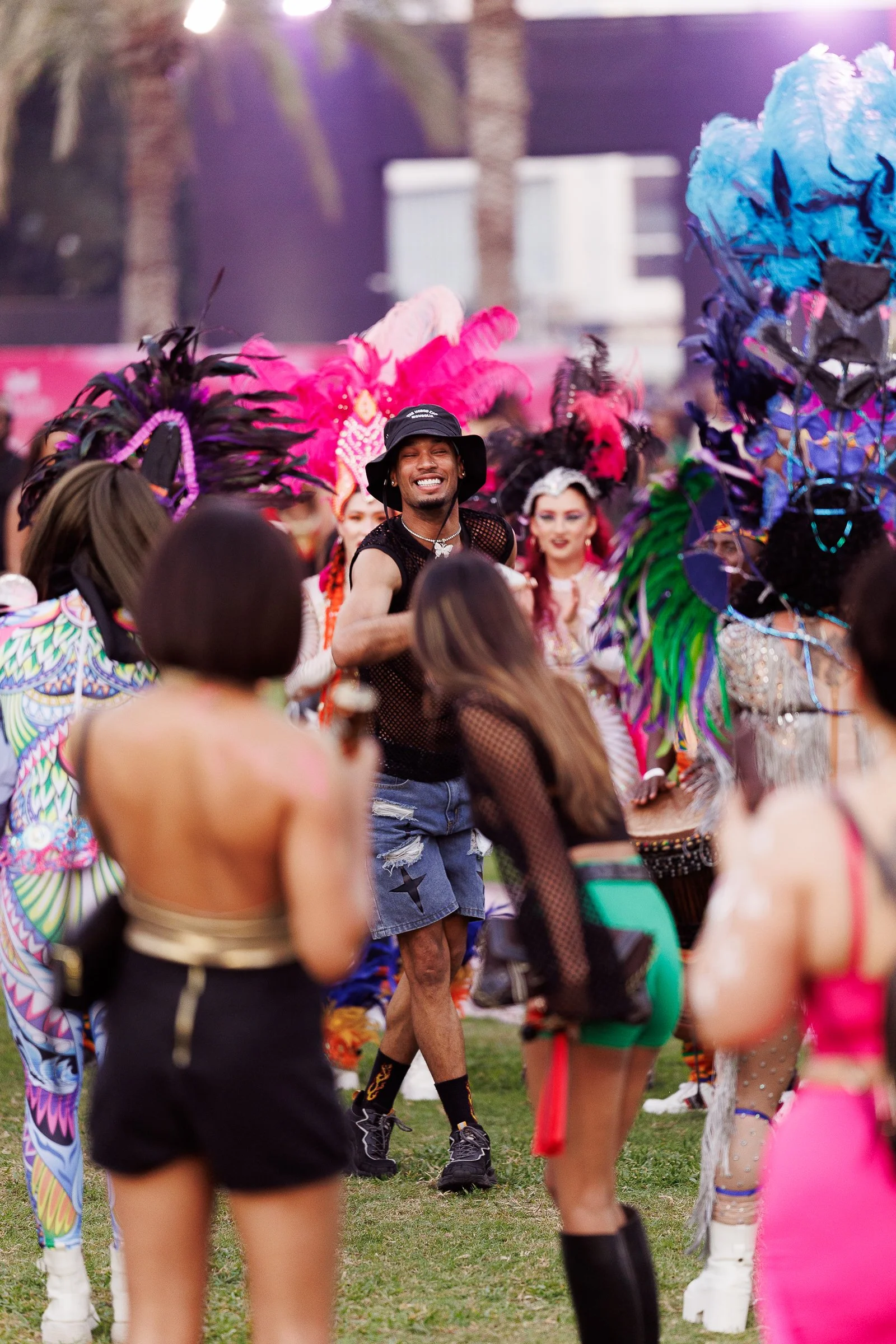 A group of people dressed in colorful costumes with feathers and bright accessories, likely participating in a festive outdoor event or parade. The focus is on a smiling man in the center wearing a black hat, mesh top, and shorts, surrounded by attendees wearing vibrant attire and headpieces.