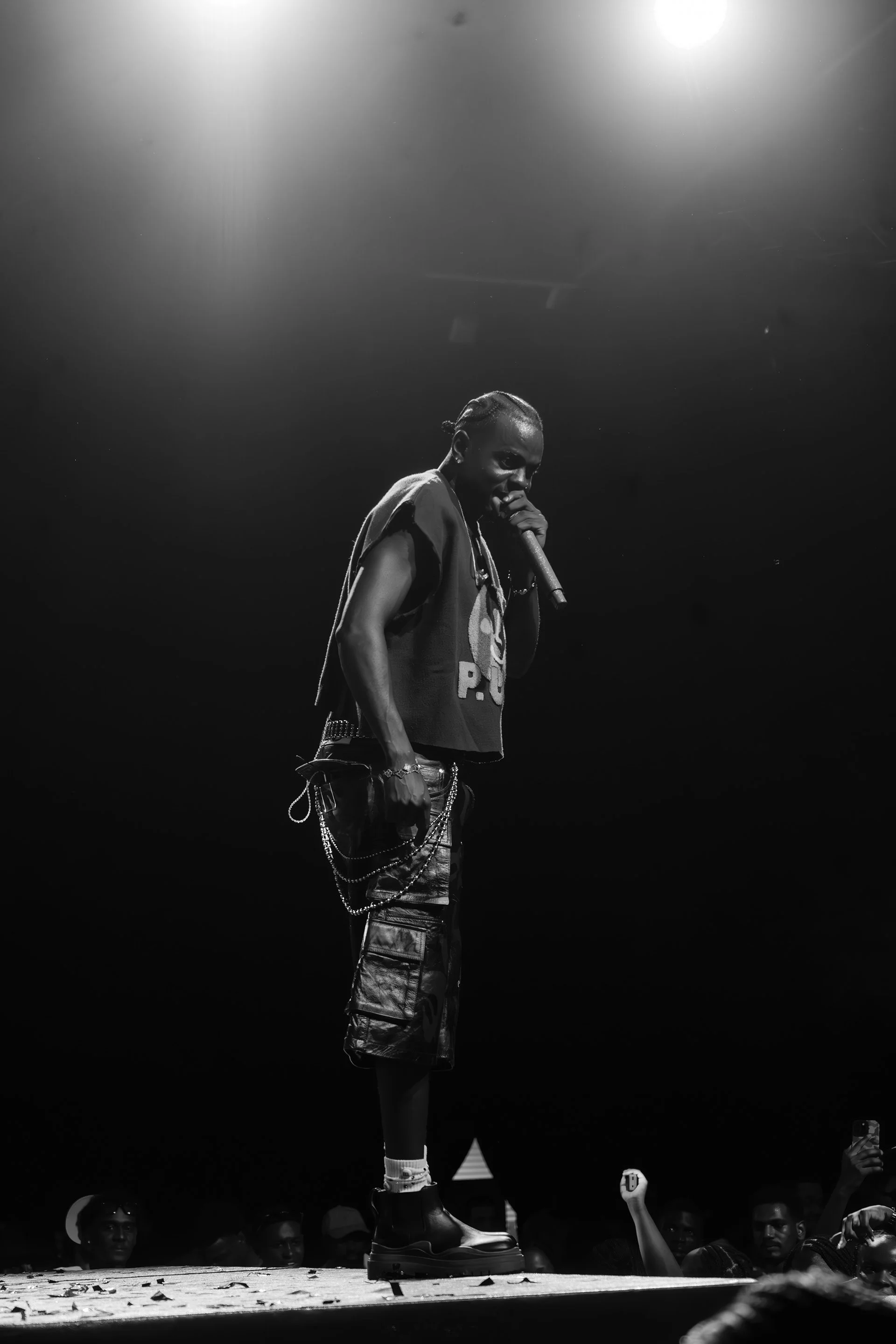 A performer at an event standing on stage, holding a microphone, wearing a sleeveless shirt, cargo shorts, and boots, with an audience visible in the background.