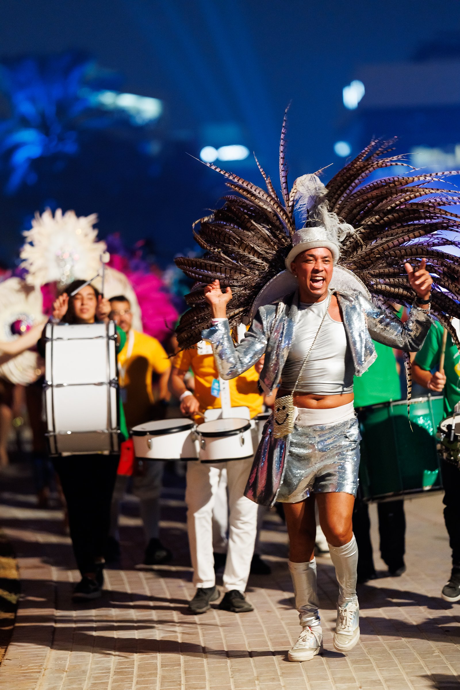 A performer dressed in a shiny silver outfit with thigh-high silver boots and a large feathered headdress, dancing and smiling during a parade or festival with a group of musicians and performers in colorful costumes in the background.
