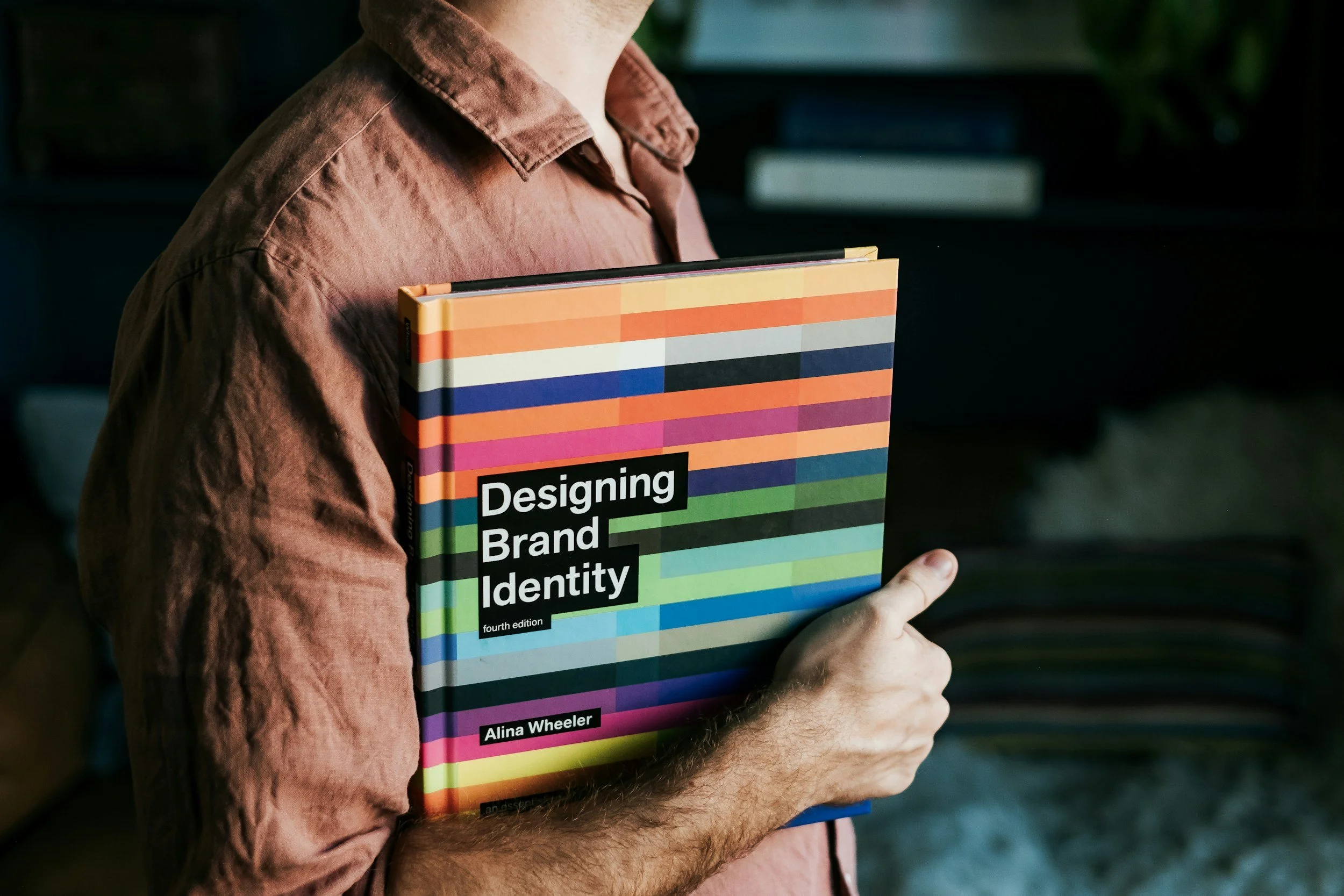 Man houdt een boek vast met een kleurrijk gestreept ontwerp en de titel "Designing Brand Identity" van Alina Wheeler.
