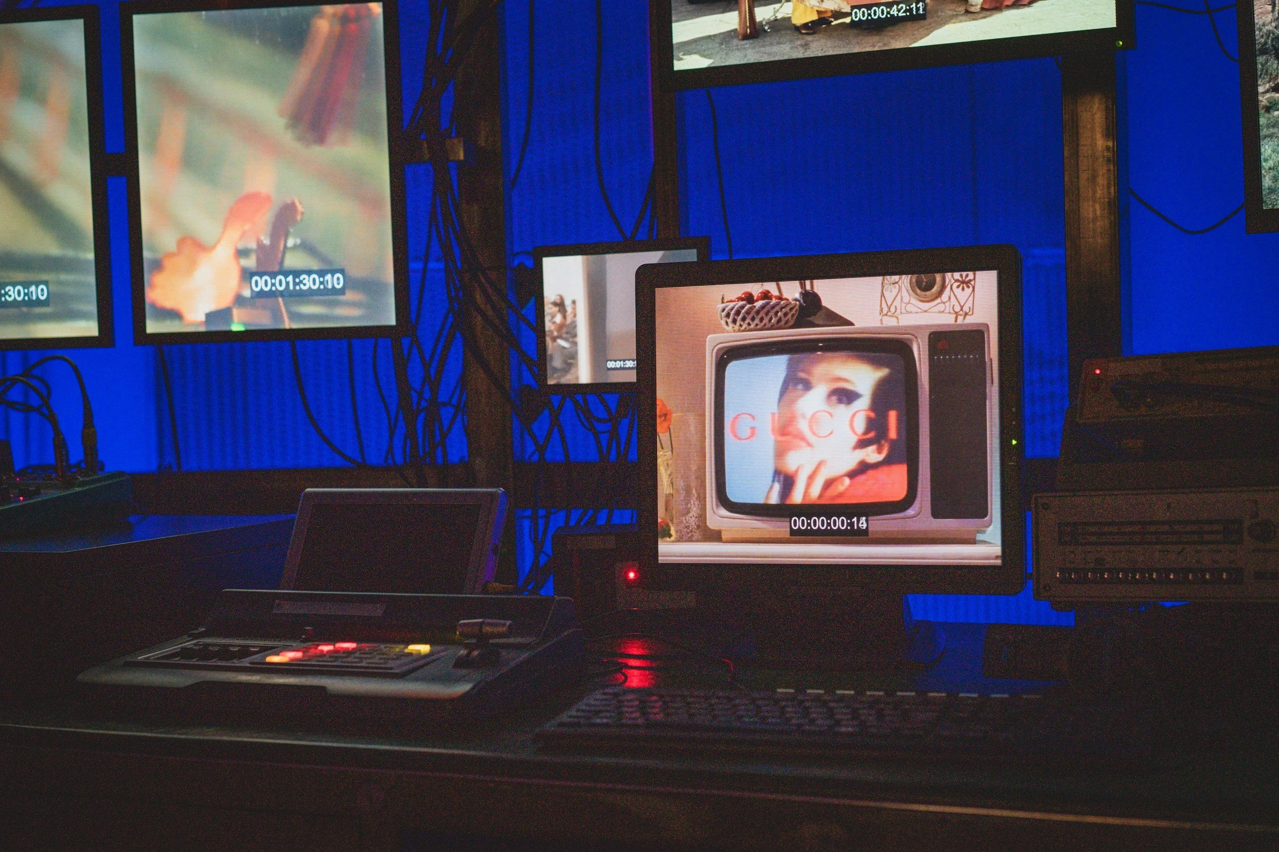 Opnamestudio met meerdere schermen, monitors en afspeelapparatuur. Eén scherm toont een close-up van een hand die een middelvinger omhoog steekt, een ander scherm toont een vrouw met make-up en een winkelwagen met de tekst 'GUCCI'.