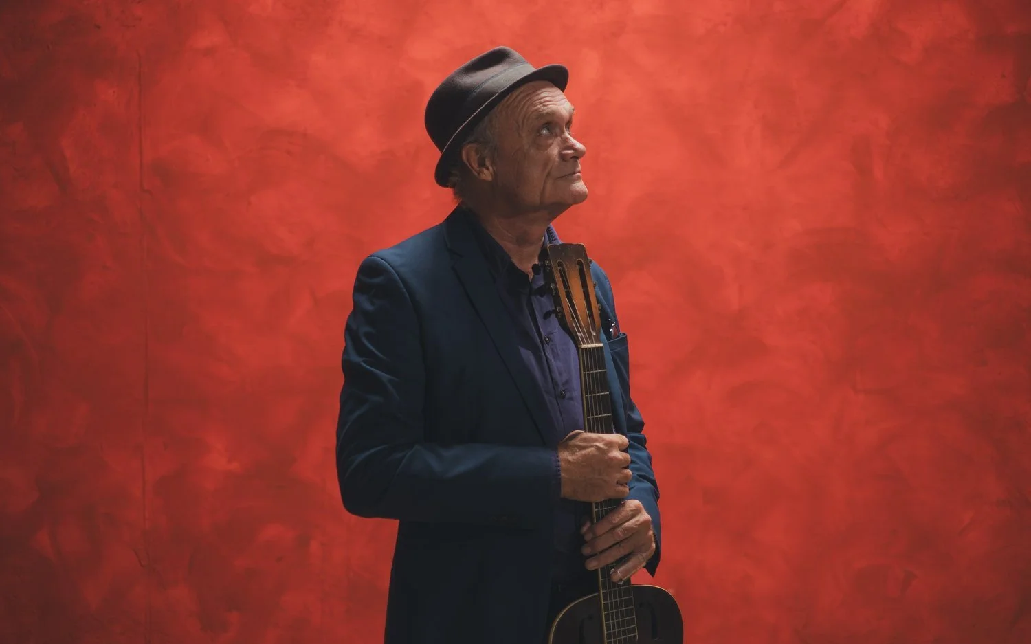 Een oudere man met een hoed, gekleed in een donkerblauw pak, houdt een gitaar vast en kijkt omhoog tegen een oranje-rode achtergrond.
