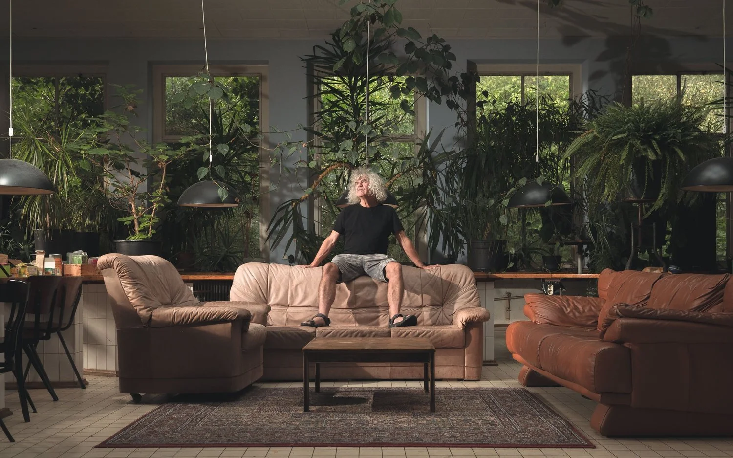 Een man met grijs, krullend haar zit op een bank in een kamer vol met grote groene planten, met grote ramen op de achtergrond. Hij draagt een zwarte T-shirt en korte broek, en lijkt ontspannen.