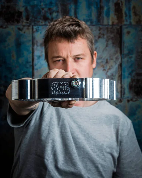 Man houdt een zware metalen slee in de hand naar de camera, met een roestige blauwe muur op de achtergrond.