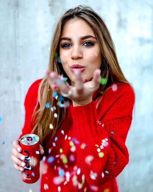 Vrouw in een rode trui blaast kleurrijk confetti in de lucht, terwijl ze een blikje frisdrank vasthoudt.