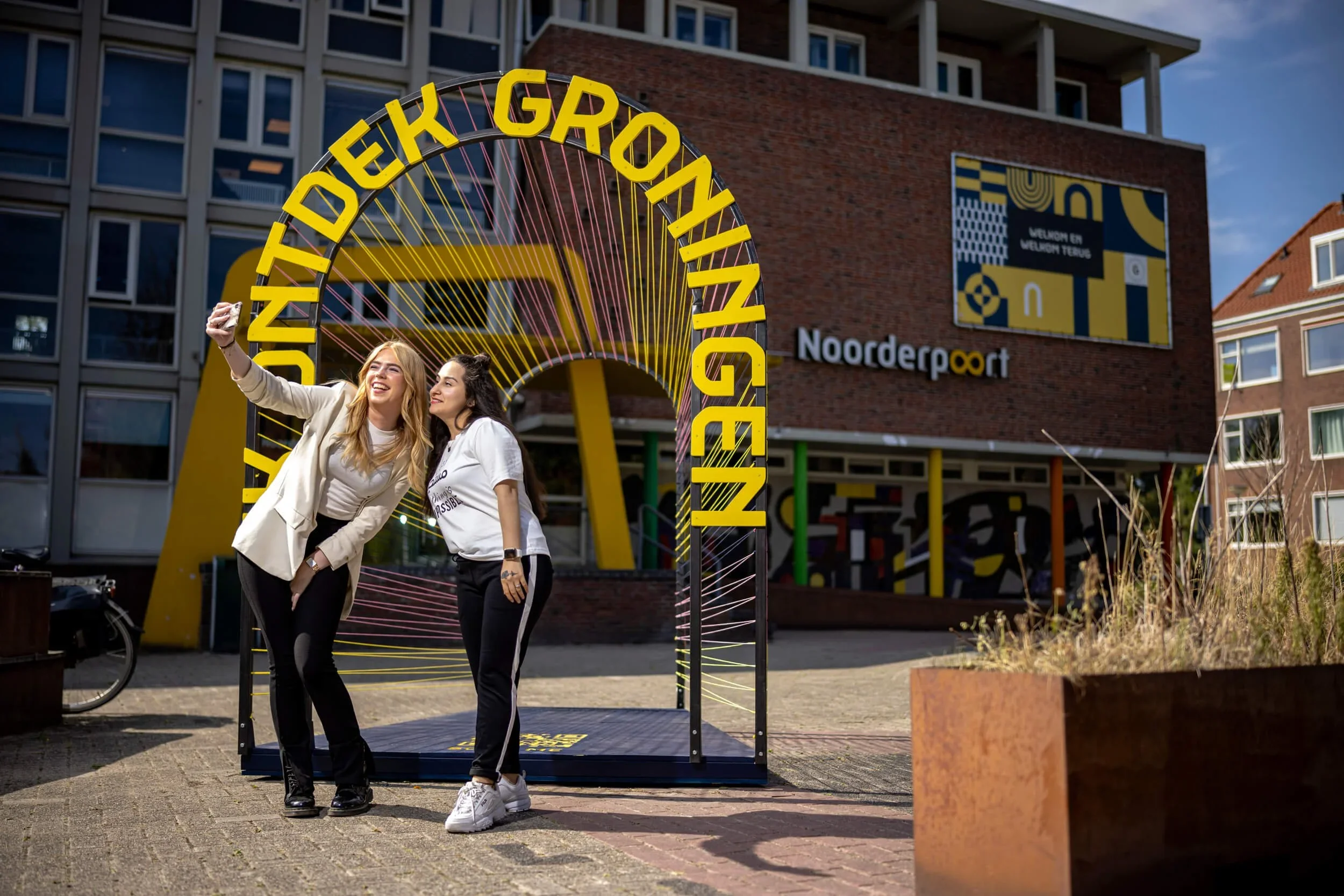 Twee vrouwen maken een selfie voor het Noorderpoort gebouw, met een kleurrijke kunstinstallatie en een boeket in de hand.