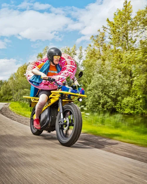 Man op een motor met zwemvliezen en een opblaasring gekleed in zwemkleding, rijdend langs een rivier met groen bomen op de achtergrond.