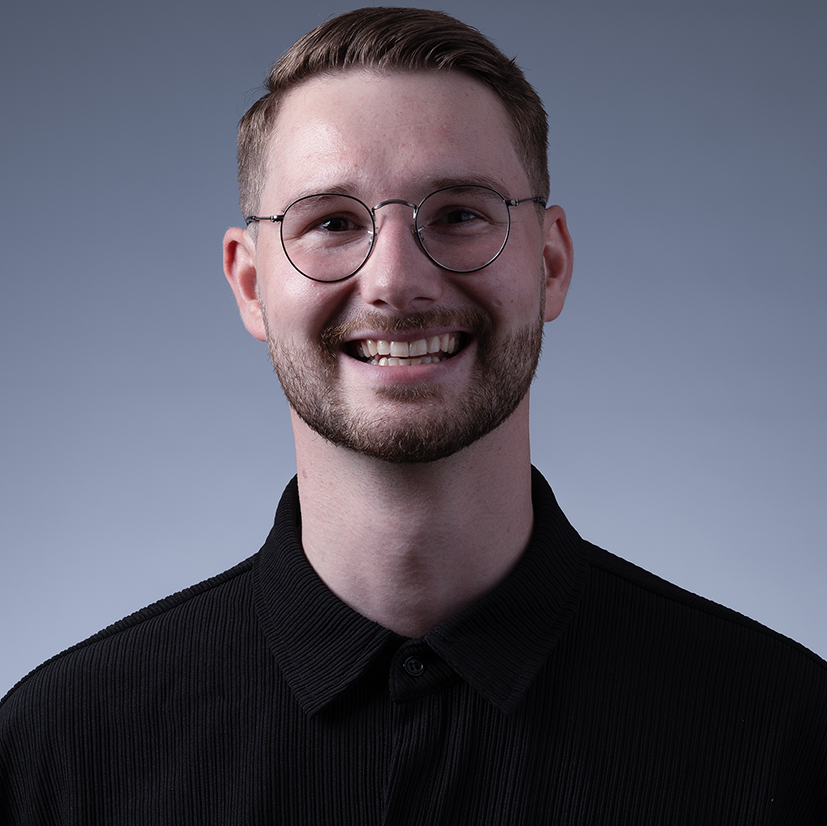 Portret van Rowdy een jonge man met bril, kort haar en een glimlach, gekleed in een zwart overhemd, tegen een effen grijze achtergrond.