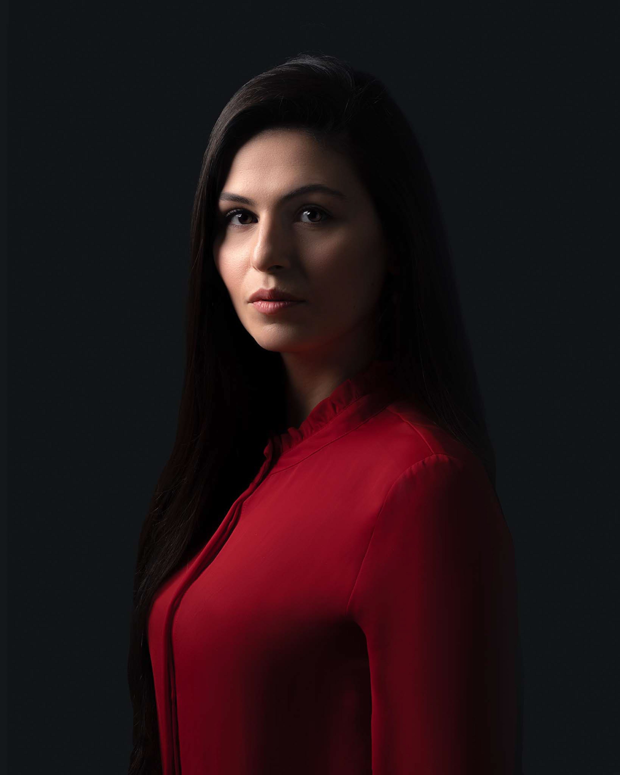 Portret van een vrouw met lang, zwart haar, gekleed in een rode blouse, tegen een donkere achtergrond.