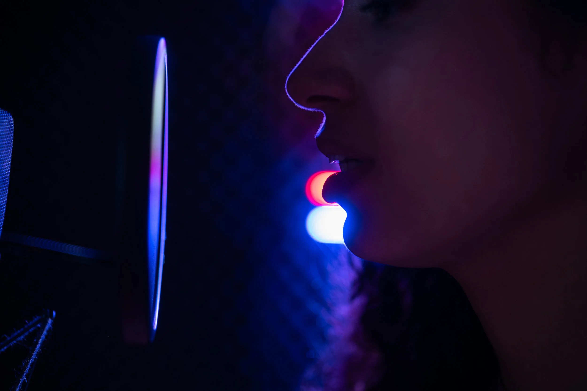 Close-up van een Kudo Voices voice-over stem die in de vocal booth spreekt in een neumann microfoon, met kleurrijk neon Kudo Media bord op de achtergrond.