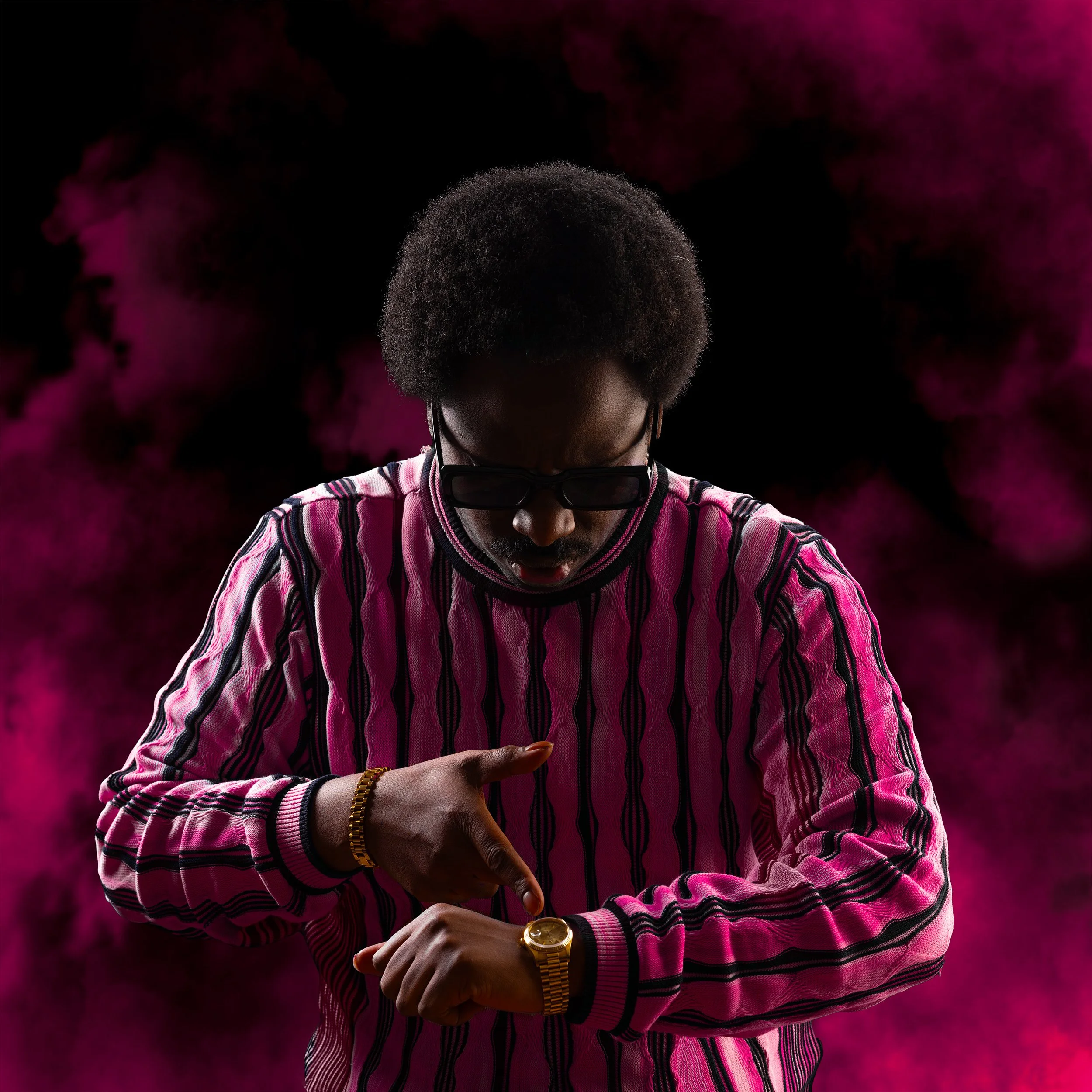 Een Foto van Mulu Glas in een roze en zwart gestreept shirt met zonnebril past zijn horloge aan, met een roze en zwart gekleurde rook op de achtergrond.