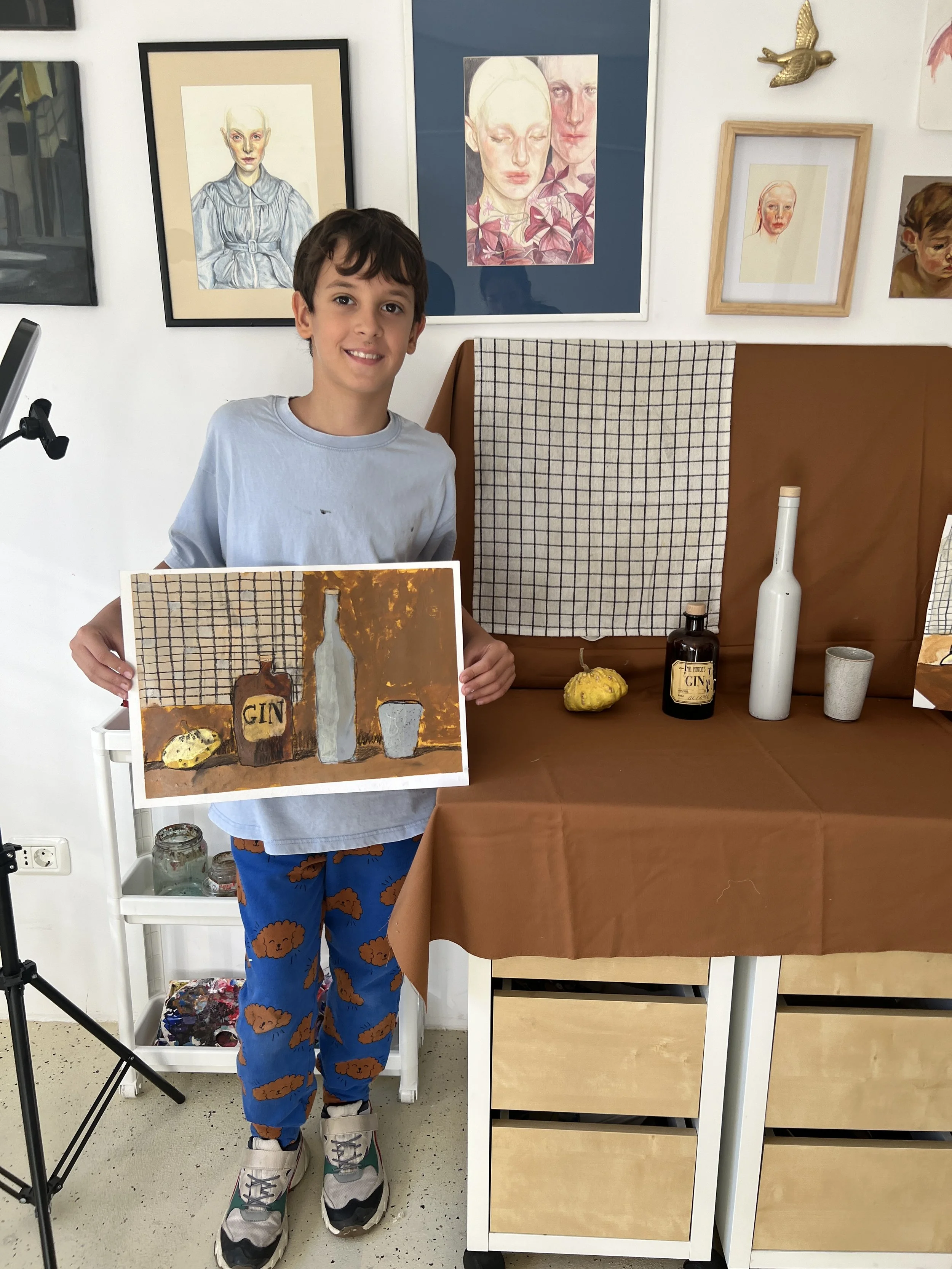 Young boy with brown hair holding a painting of a still life with a bottle, a cup, and a lemon in front of a brown table with bottles and a pumpkin, in an art studio with framed portrait paintings on the wall.