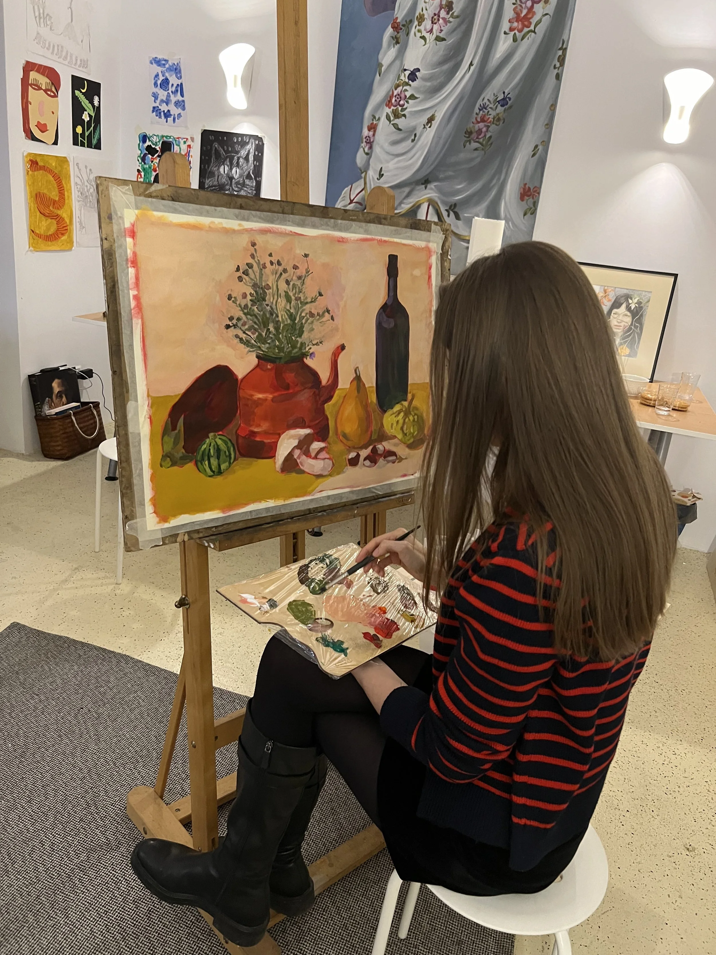 A woman with long brown hair wearing a black and red striped sweater, black skirt, and black boots sits on a white stool and paints a still life on a canvas in an art studio.