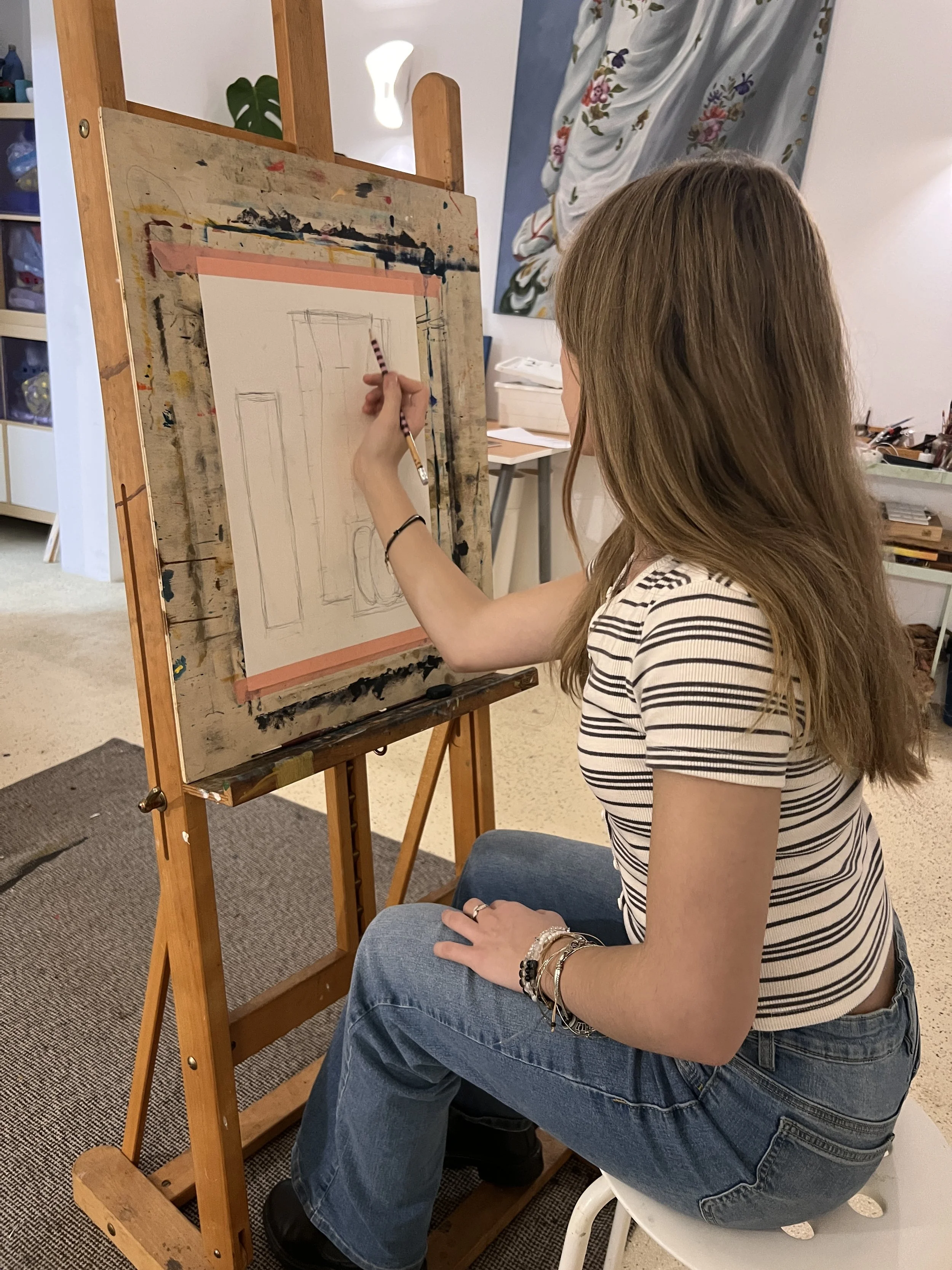 A young woman with long hair, wearing a striped t-shirt and jeans, is sitting on a white stool, drawing geometric shapes on a canvas set on an easel in an art studio.