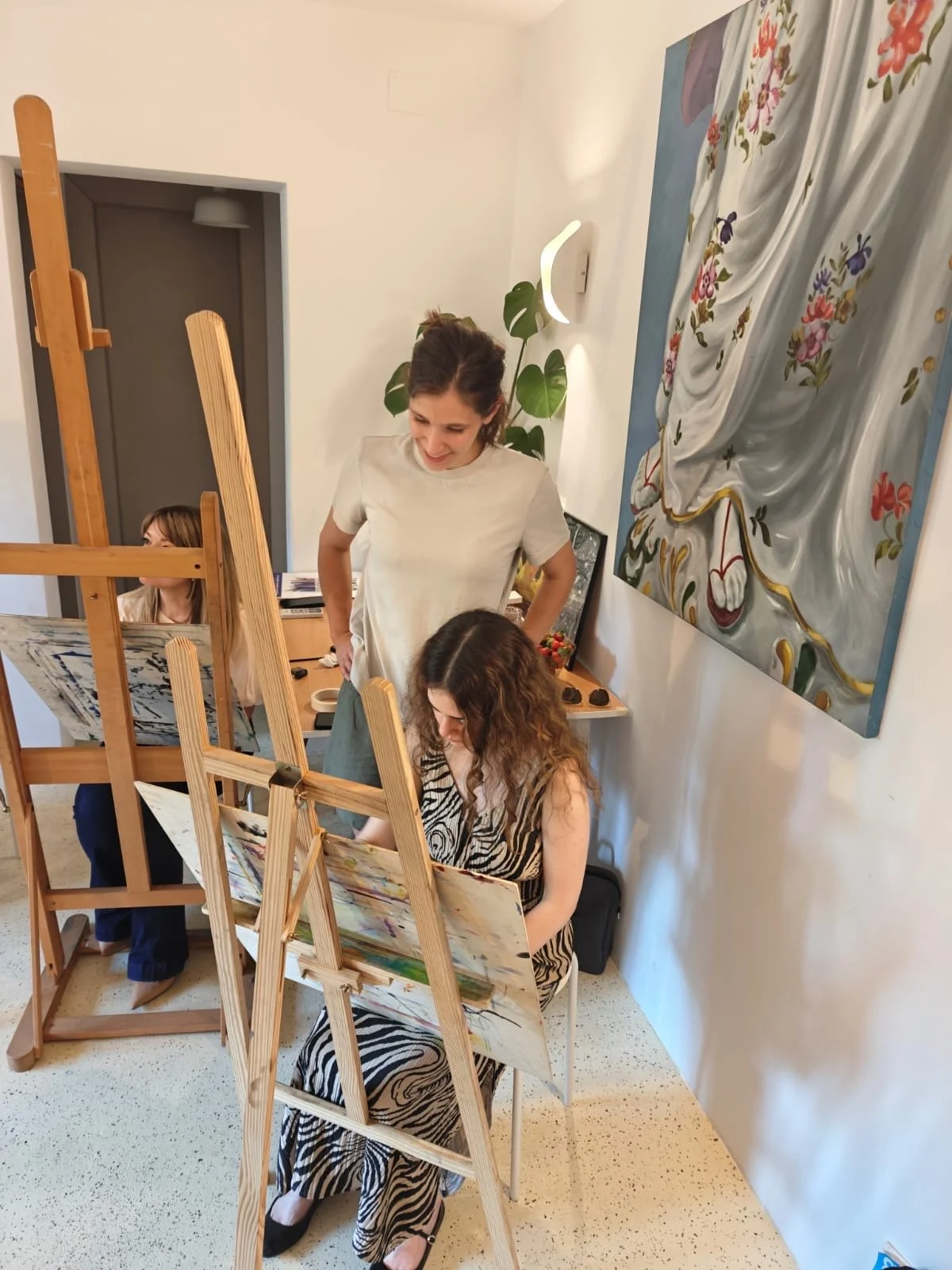 Two women, one with long wavy hair wearing a zebra print dress and sitting on a white chair, painting on a canvas at an art class, with another woman with curly hair and a white shirt standing nearby, observing. There are art easels, paintings, and a