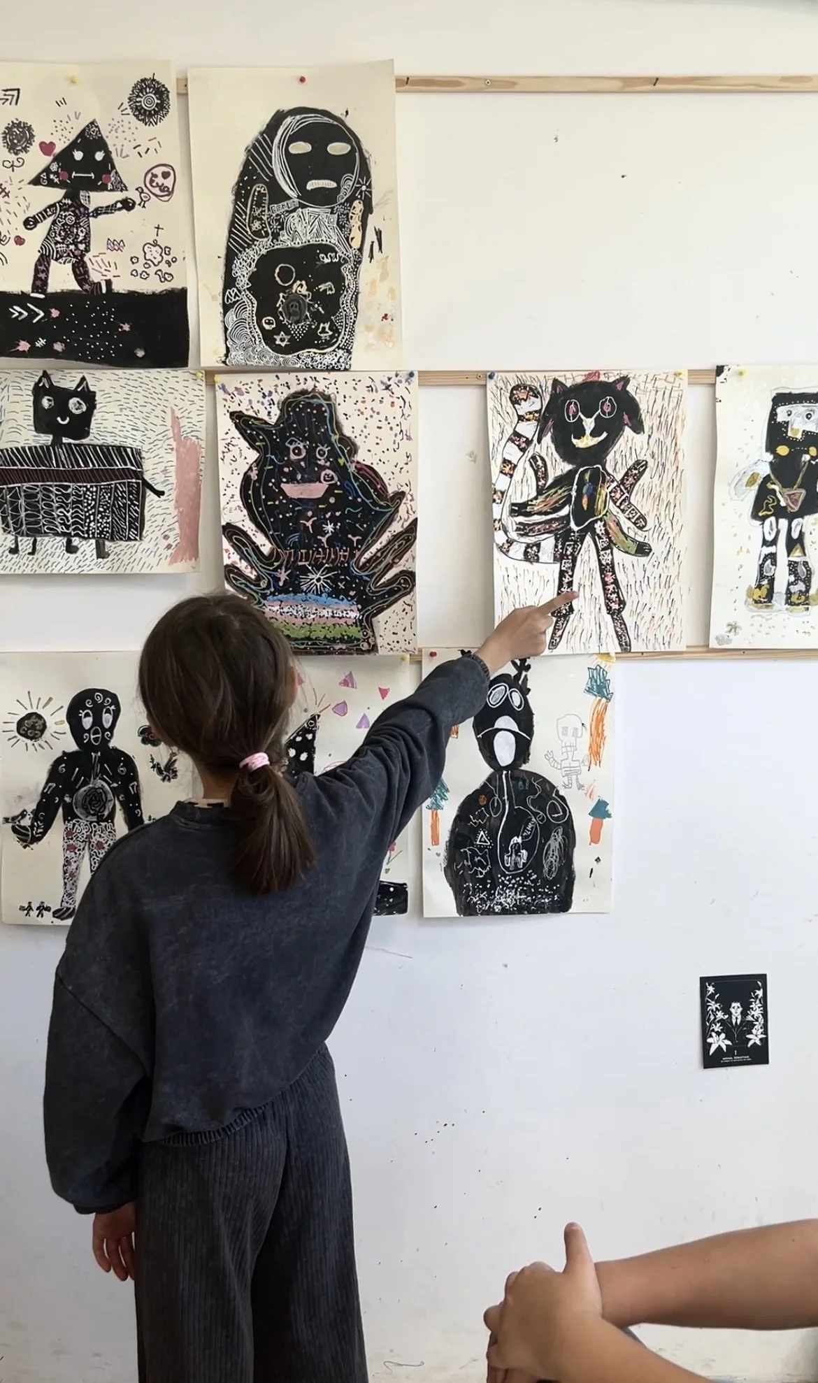 A young girl with a ponytail pointing at artwork on a wall in an art gallery. The wall features various black and white drawings and paintings of abstract, animal-like, and humanoid figures.
